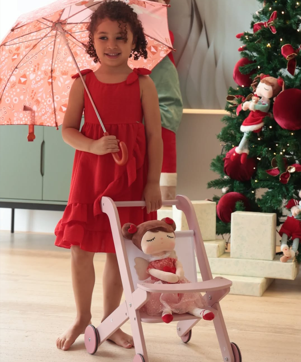 Menina veste vestido vermelho de algodão e segura guarda-chuva rosa para brincar livre empurrando carrinho com boneca.