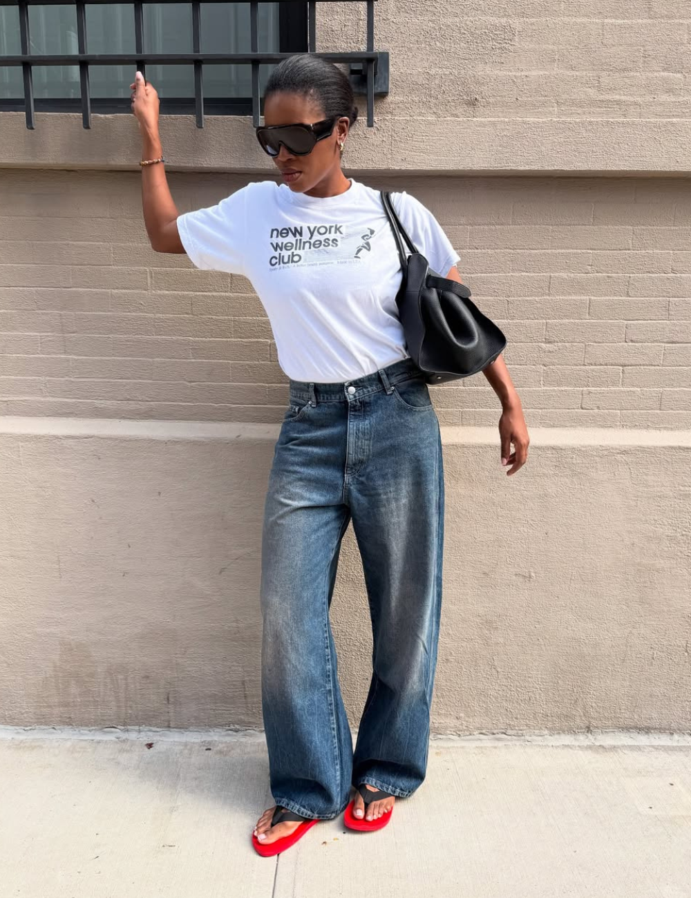 Modelo posa com calça jeans boyfriend azul de lavagem escura, camiseta branca, bolsa preta e chinelos vermelhos.