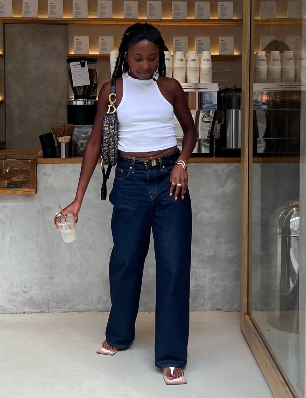 Modelo posa com calça jeans boyfriend azul escuro de modelagem ampla, combinada a regata branca justa e sandálias quadradas.