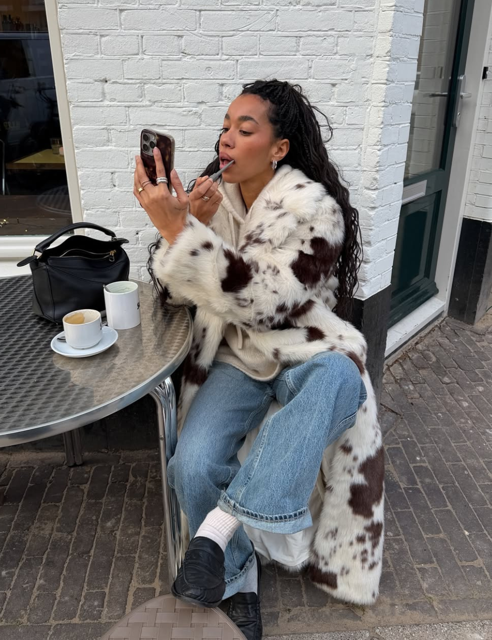 Modelo sentada aplica batom olhando no celular, veste calça jeans boyfriend azul clara, casaco peludo branco com manchas.