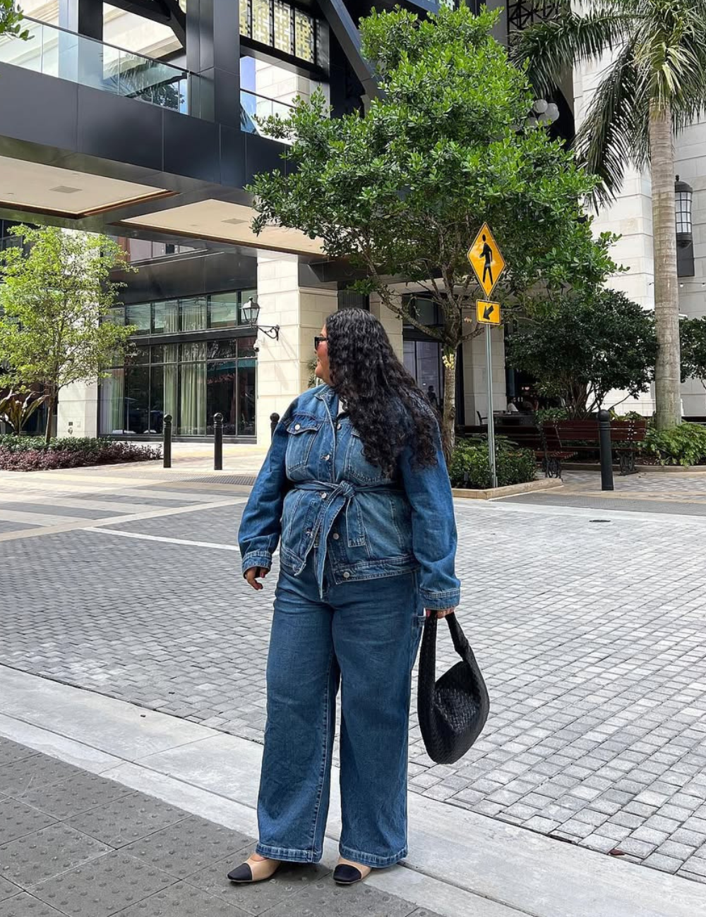 Modelo posa na rua com calça jeans boyfriend azul de corte largo, jaqueta jeans amarrada na cintura e bolsa preta de mão.