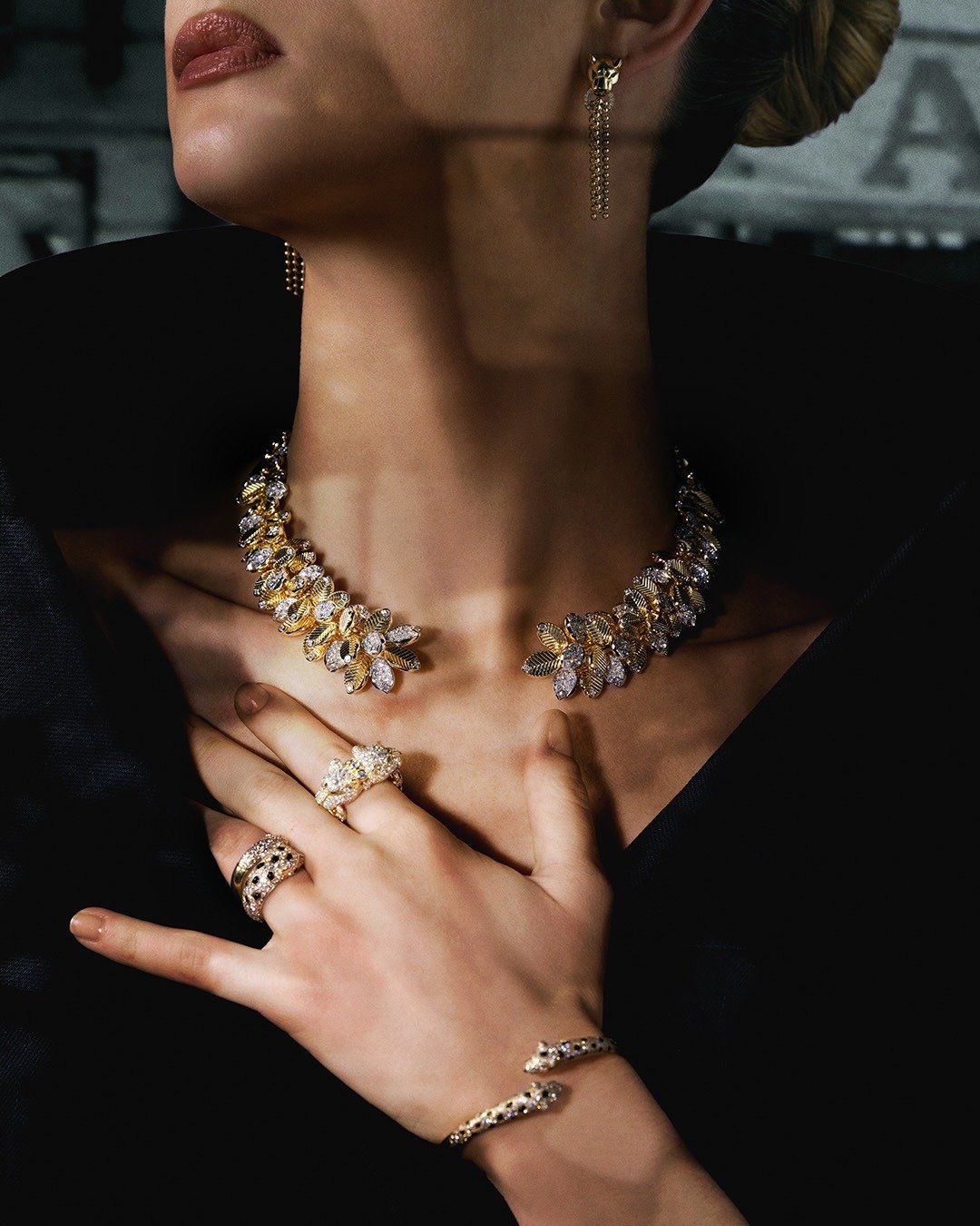 Modelo posa com postura elegante usando colar, anéis e pulseira dourados com pedras brilhantes sobre vestido preto,