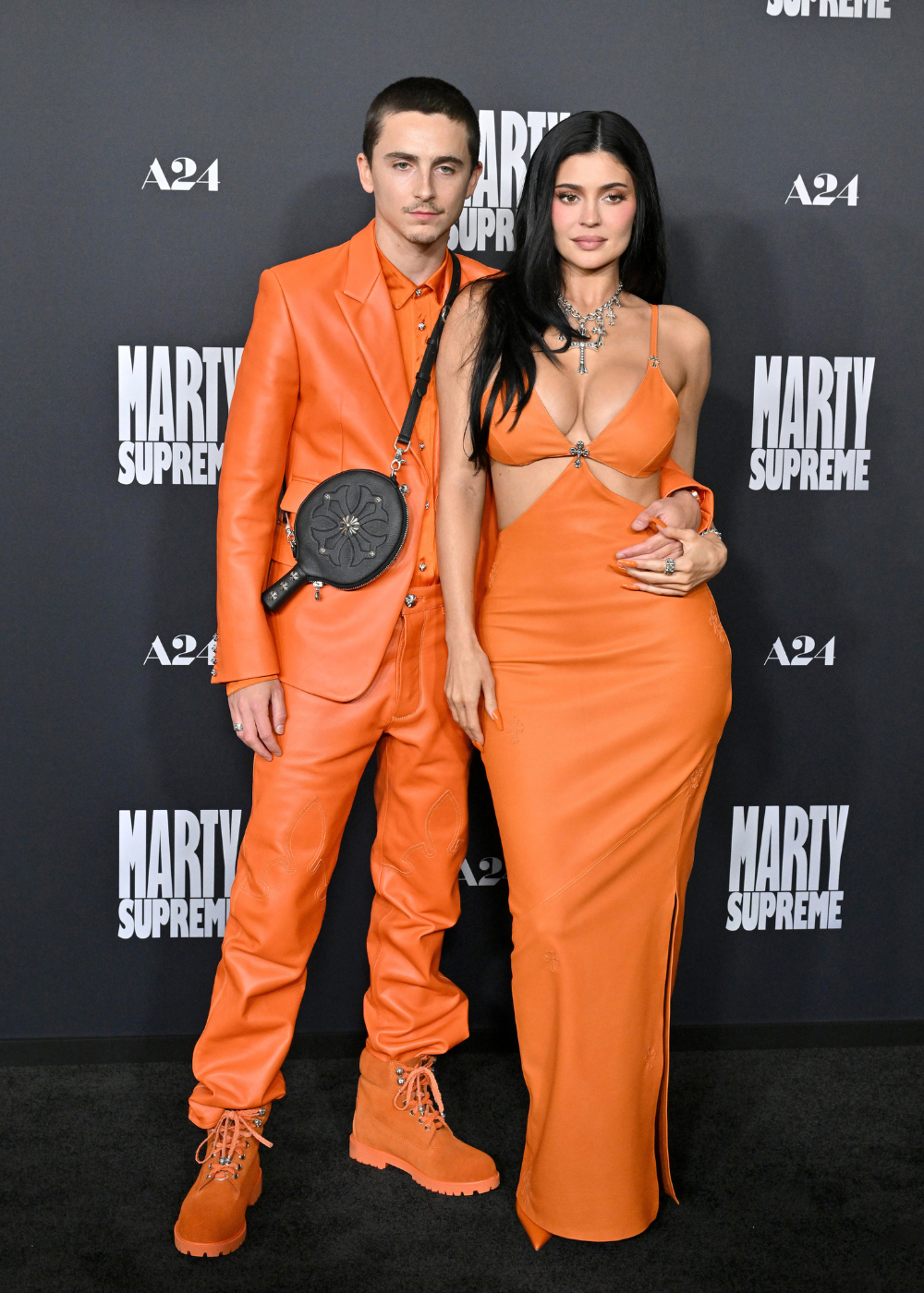 Casal de Hollywood posa junto em look monocromático laranja vibrante com couro e vestido recortado, exalando estilo moderno.
