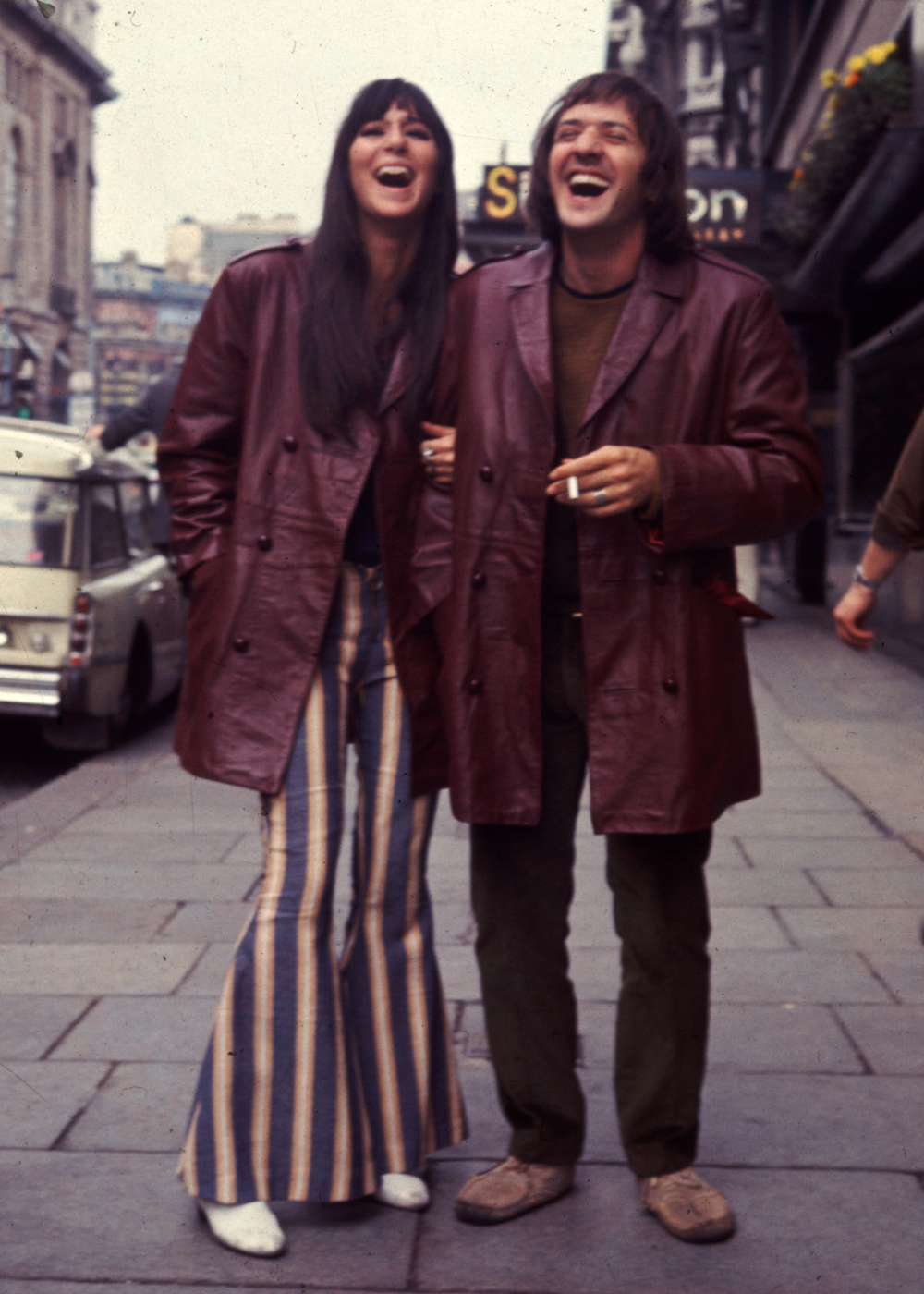 Casal de Hollywood posa rindo na rua com casacos de couro vinho, calça flare listrada e clima leve e descontraído.