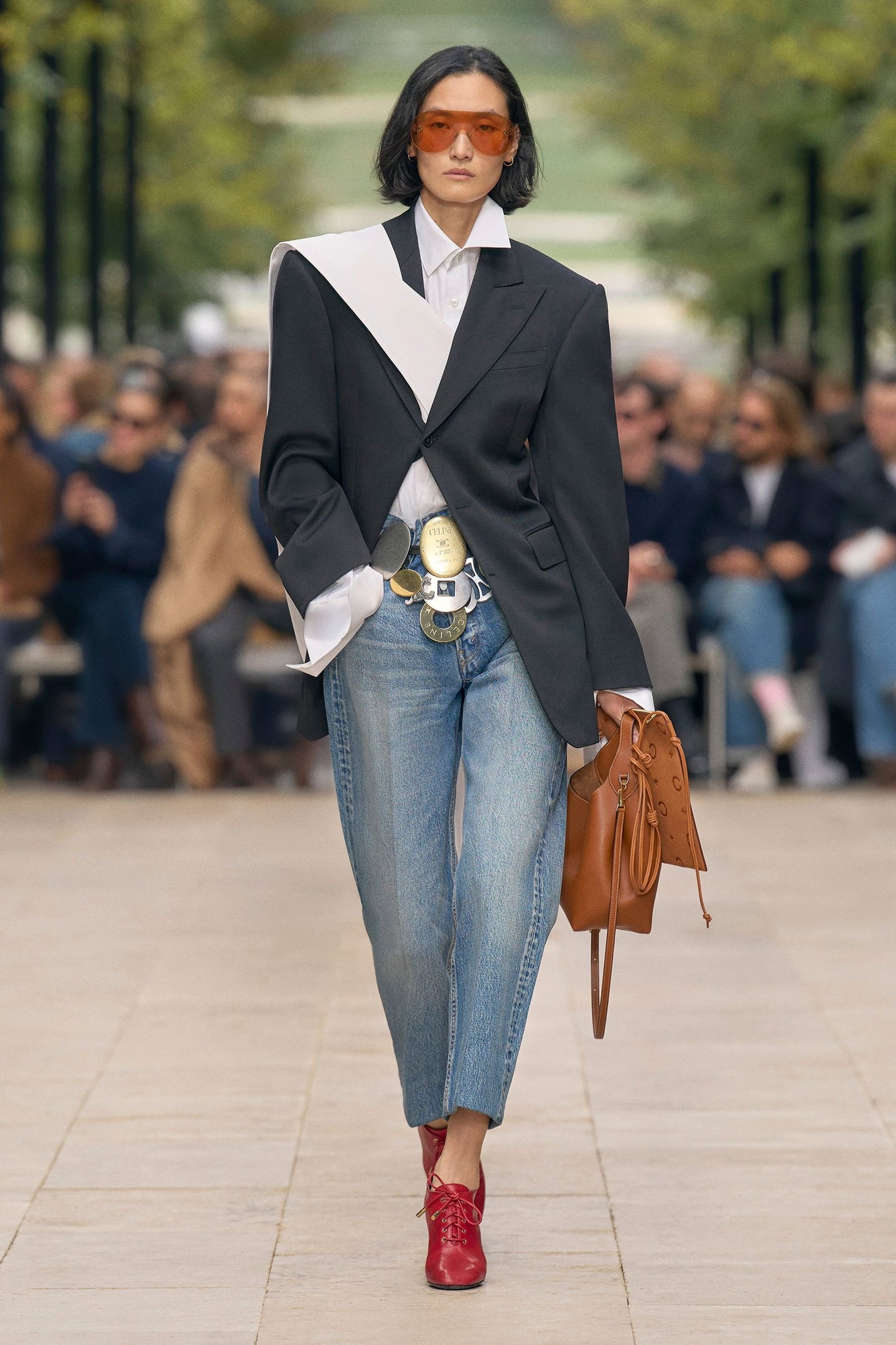 Modelo caminha confiante com calça barrel jeans azul clara de corte reto, blazer preto oversized, camisa branca e sapatos