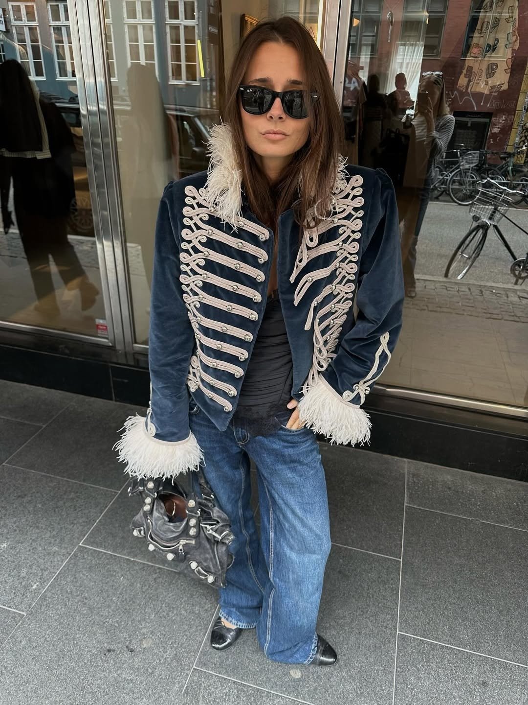 Modelo posa com jaqueta de veludo azul marinho com nó chinês branco e detalhes em plumas, combinada a jeans e bolsa cinza,