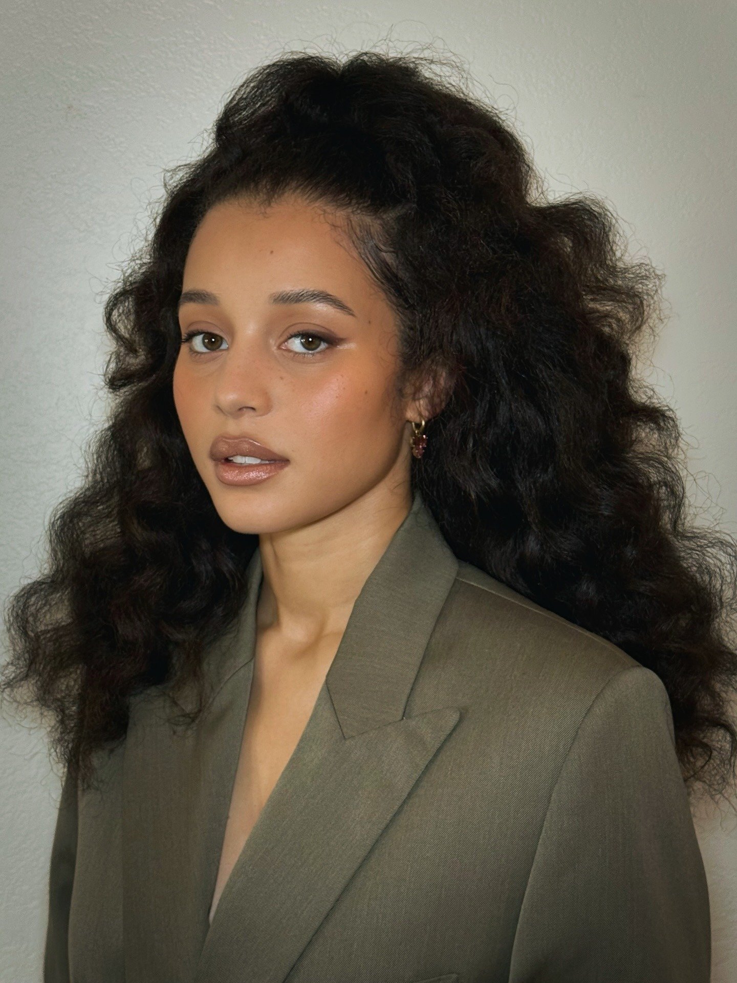 Chase Infiniti posa com blazer oversized verde-oliva de tecido estruturado, cabelo volumoso e maquiagem natural