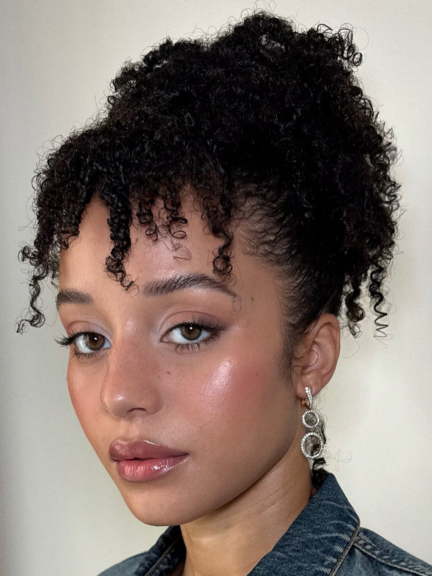 Chase Infiniti exibe maquiagem glow natural com gloss rosado, brincos prateados elegantes e jaqueta jeans