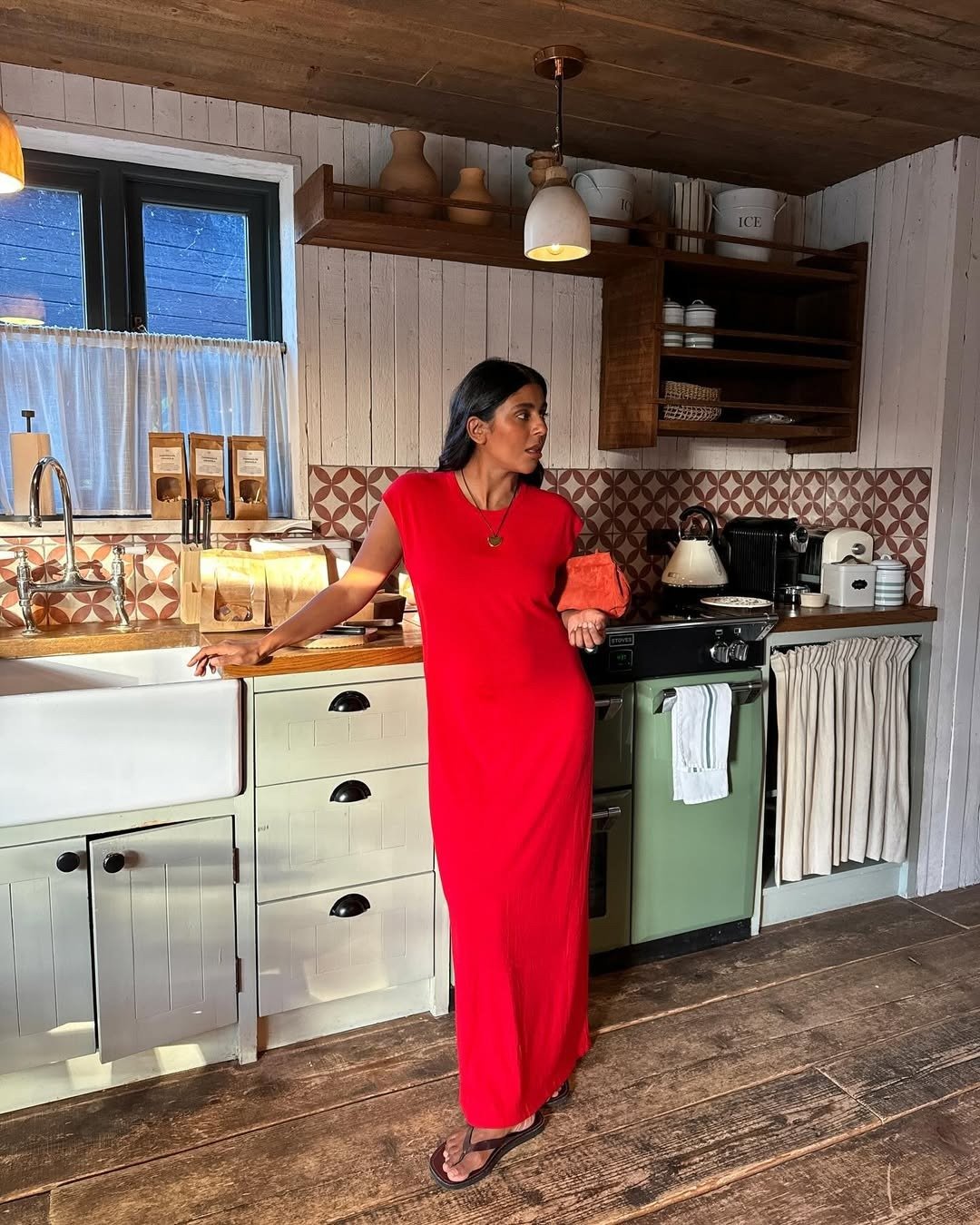 Modelo posa em cozinha rústica com vestido longo vermelho de malha e chinelo de dedo chique preto, equilibrando conforto