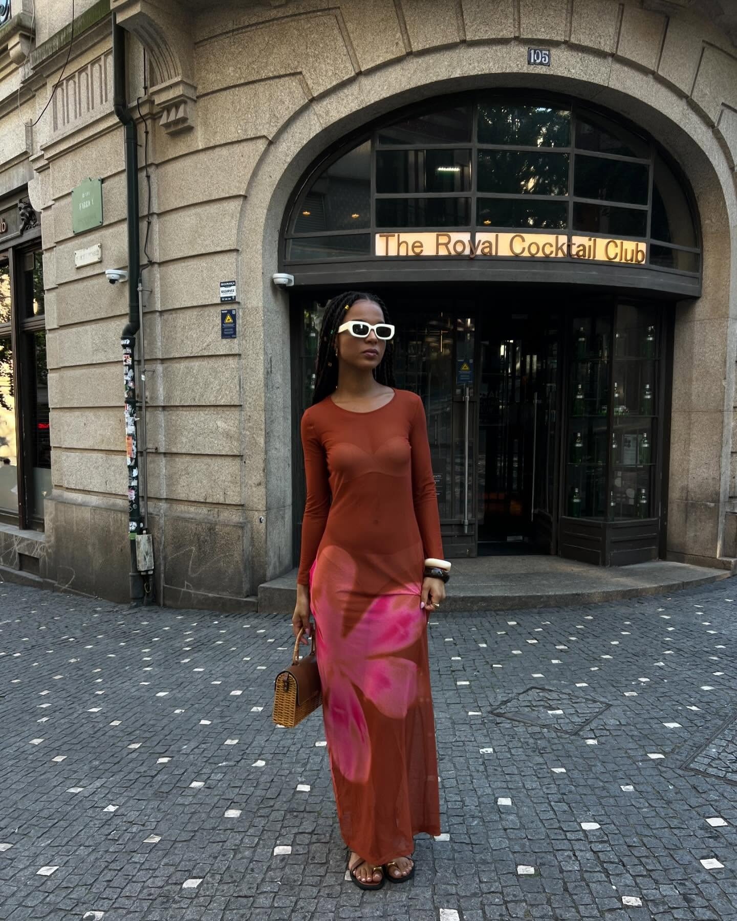 Modelo posa confiante com vestido longo justo marrom com estampa rosa, acessórios em branco e marrom, e chinelo de dedo