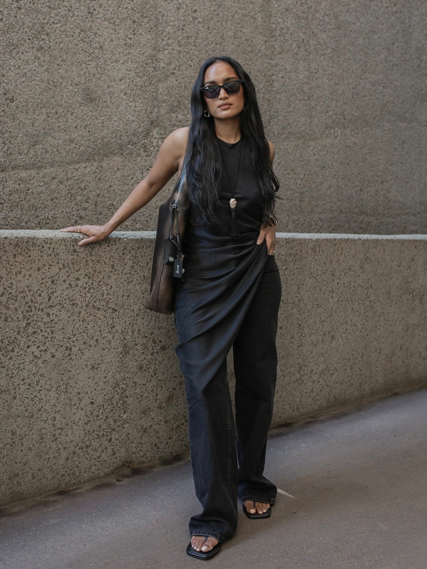 Mulher posa com vestido preto drapeado, calça jeans escura e chinelo de dedo chique preto com bico quadrado, em clima urbano
