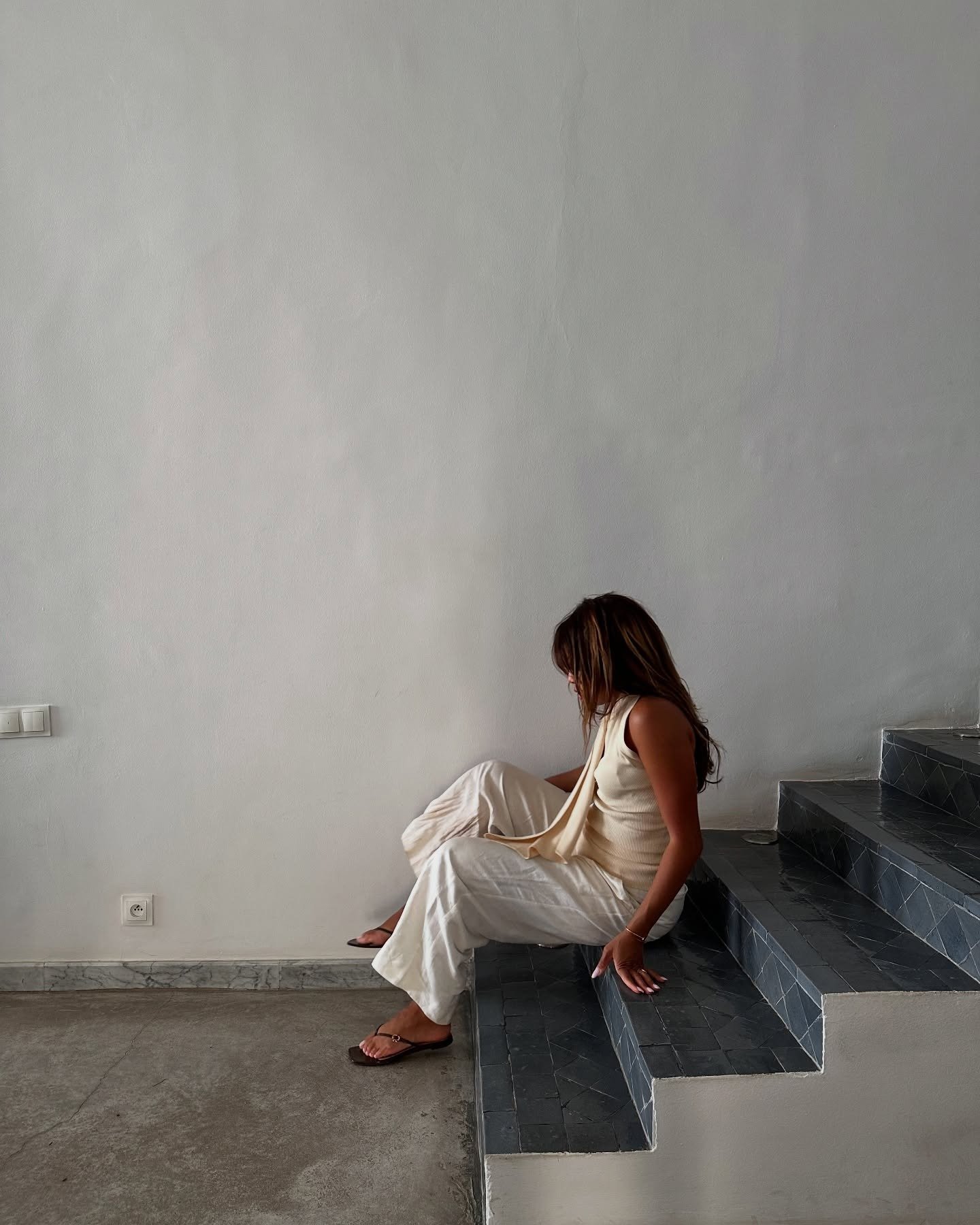 Mulher sentada em escada de azulejos escuros usa chinelo de dedo chique marrom com detalhe dourado, calça e regata bege