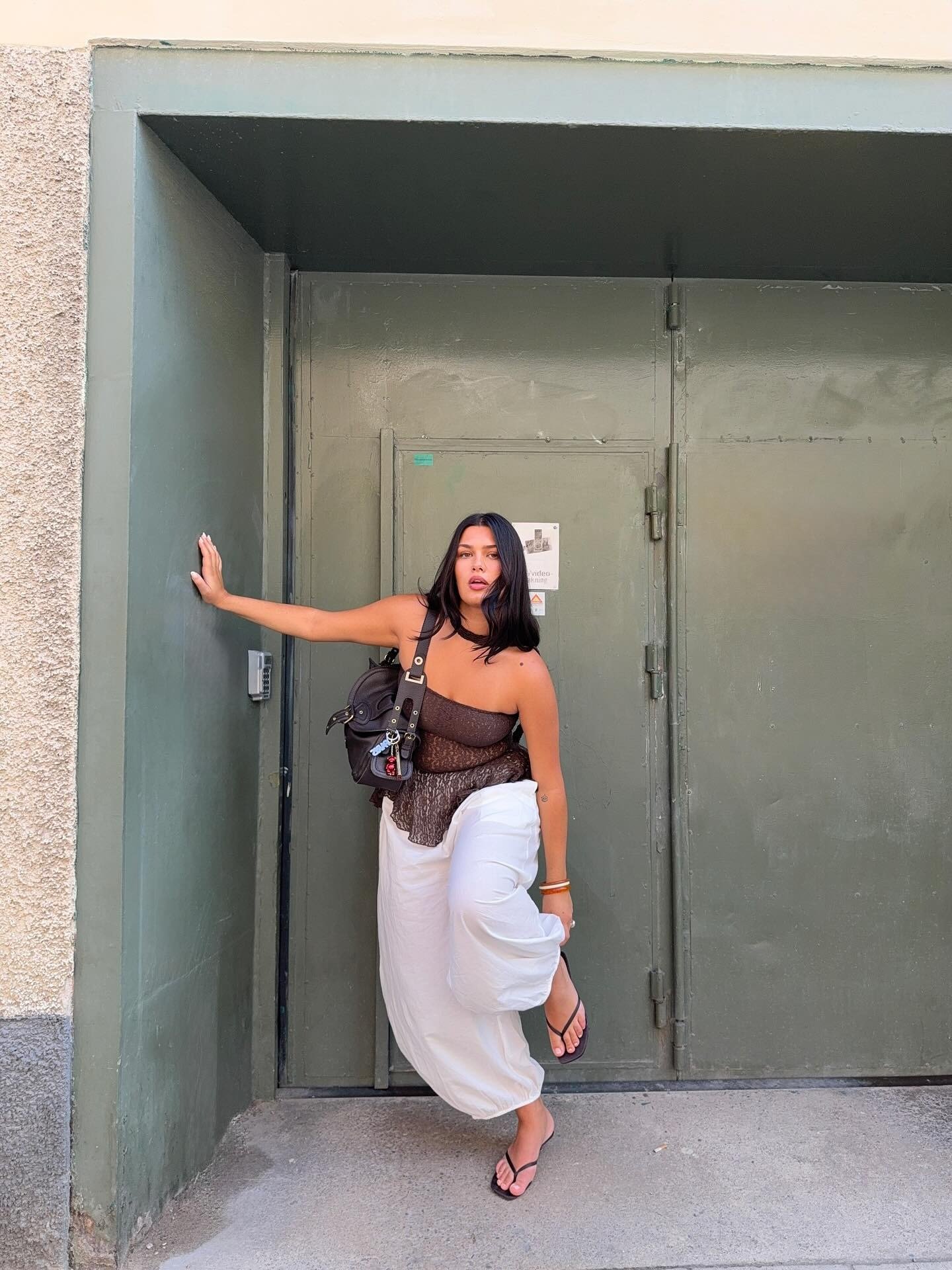 Modelo posa com postura confiante ao apoiar braço na parede, usando chinelo de salto alto preto, blusa marrom transparente