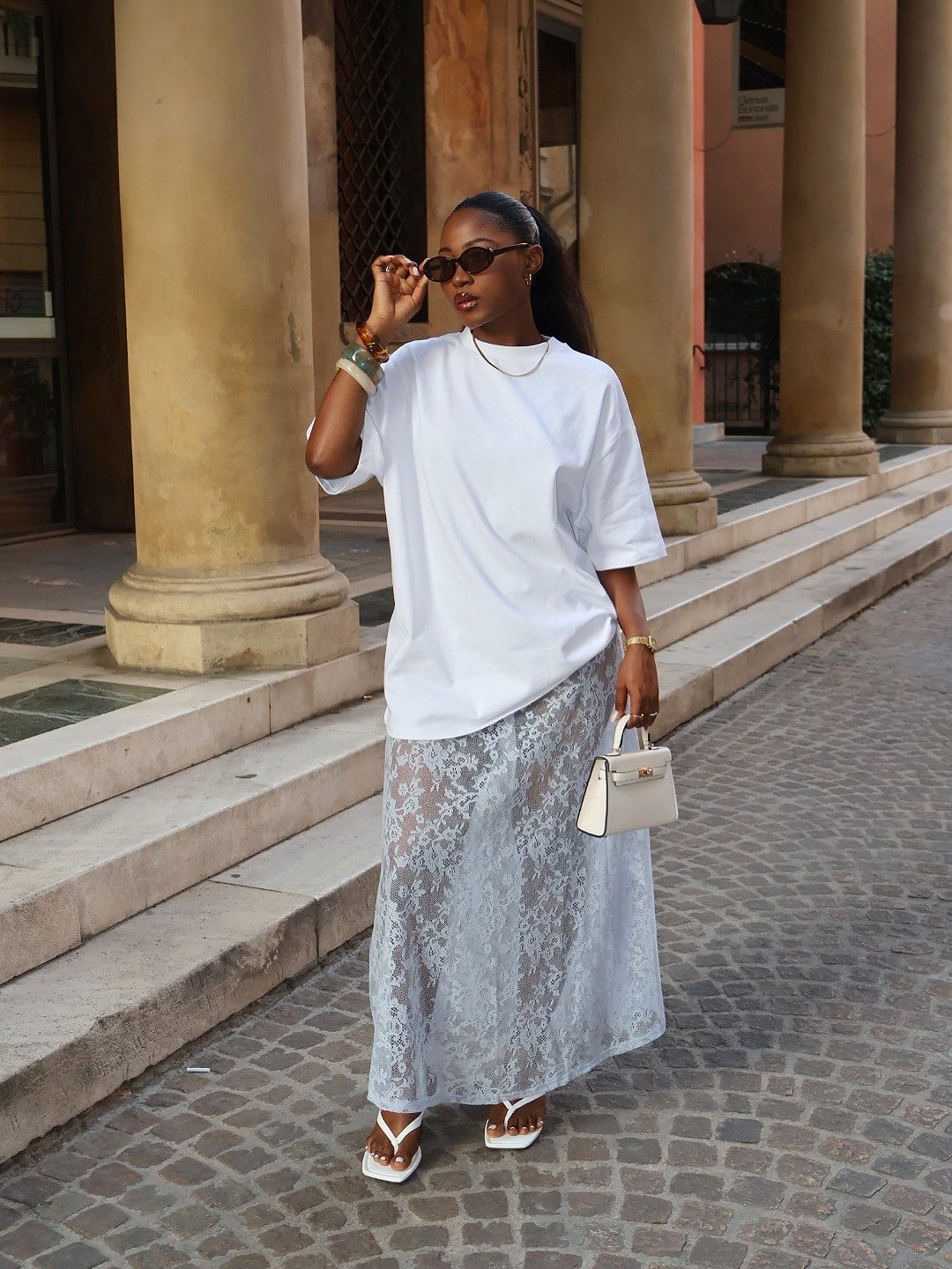 Modelo posa confiante em chinelo de salto alto branco com tiras finas, combinando saia longa rendada e camiseta oversized