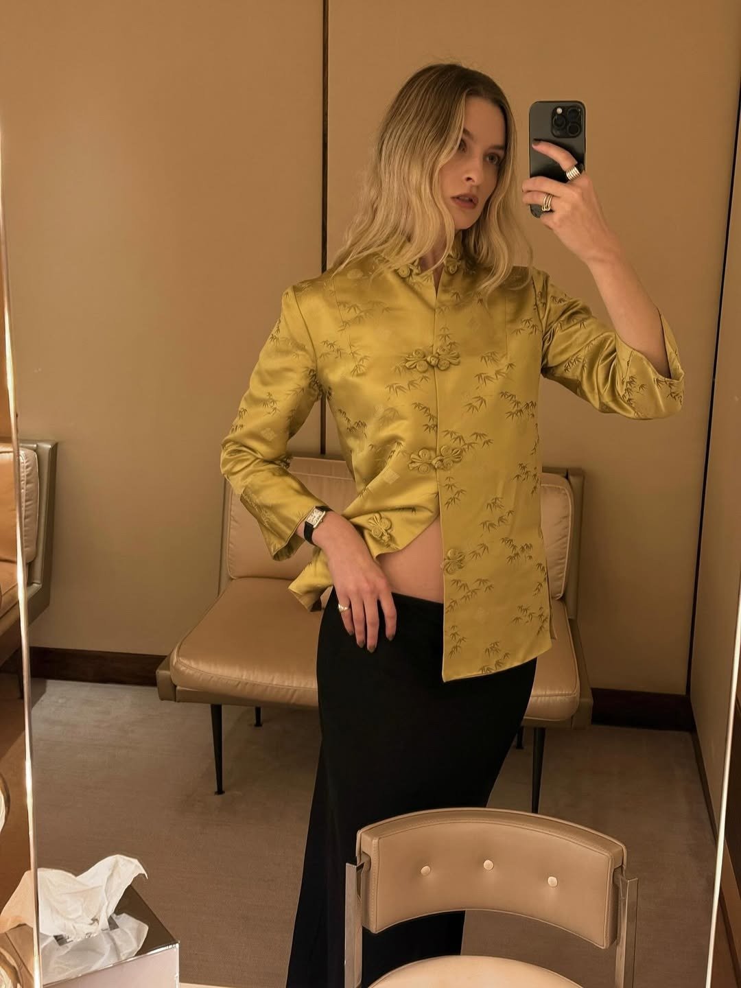Modelo posa para selfie vestindo blazer dourado de seda com nó chinês e saia preta longa, em ambiente neutro e elegante.