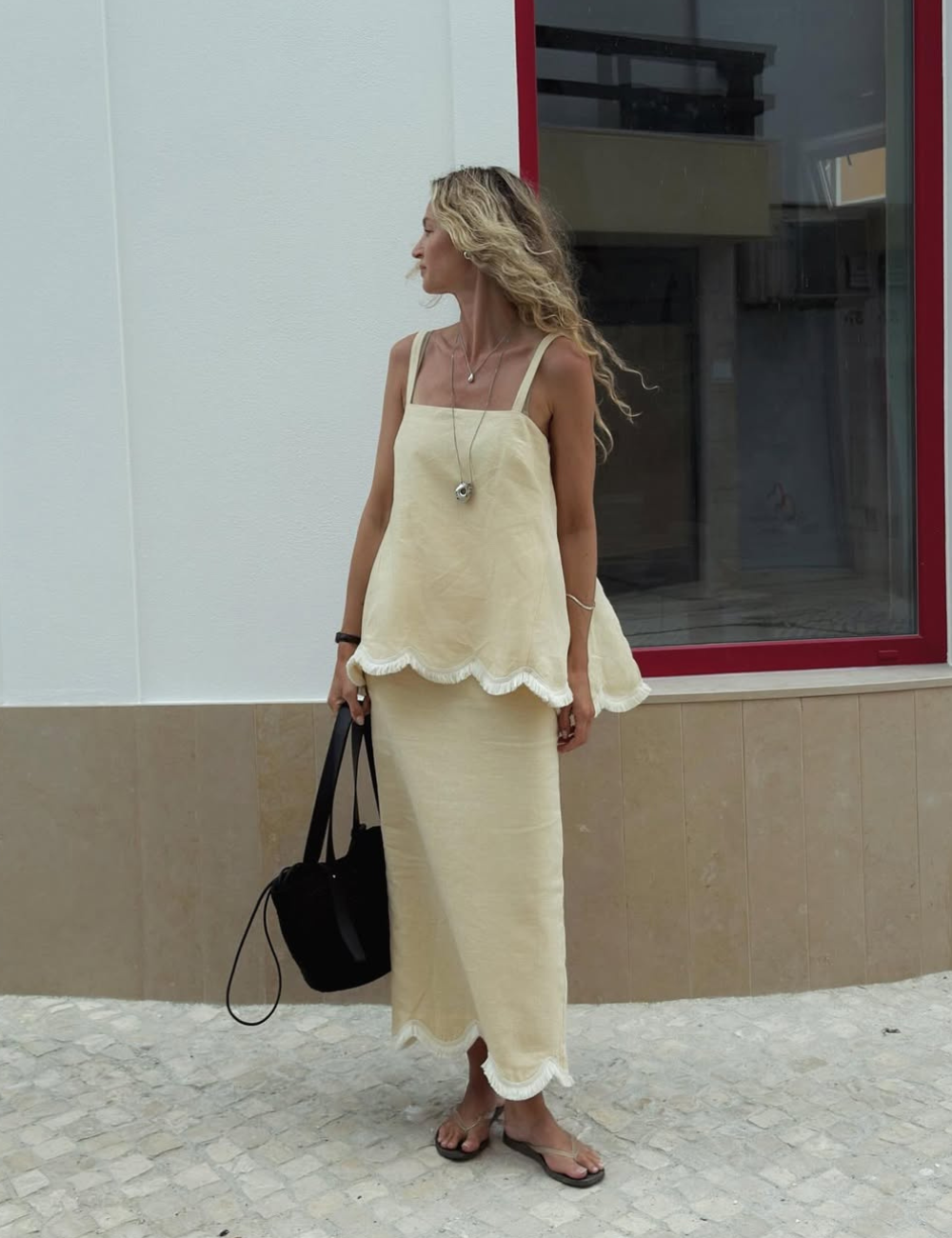 Se vestir bem com vestido de linho bege de modelagem solta e barra ondulada, combinando colares delicados e bolsa preta.