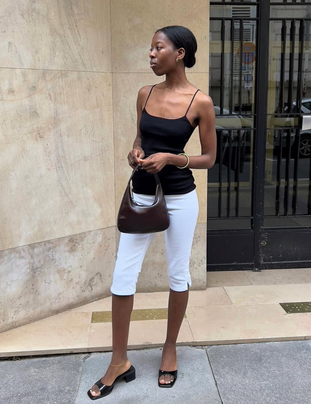 Modelo posa de perfil com regata preta de alças finas, bermuda branca justa, sandálias pretas de salto baixo e bolsa marrom.