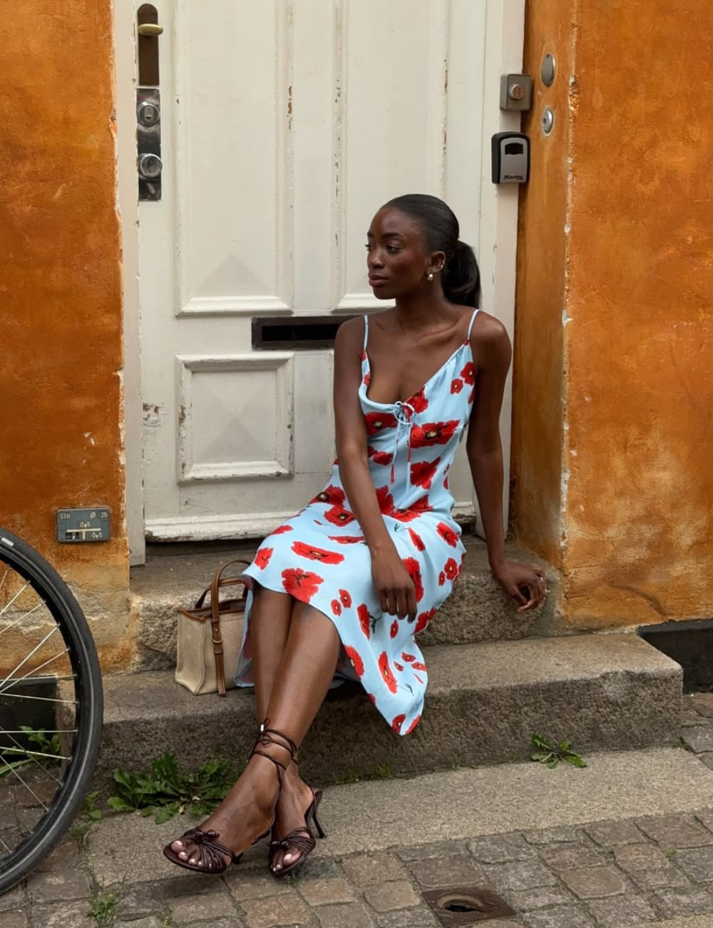 Mulher senta com postura elegante em vestido azul claro de tecido leve com estampa vermelha floral, sandálias de tiras finas.