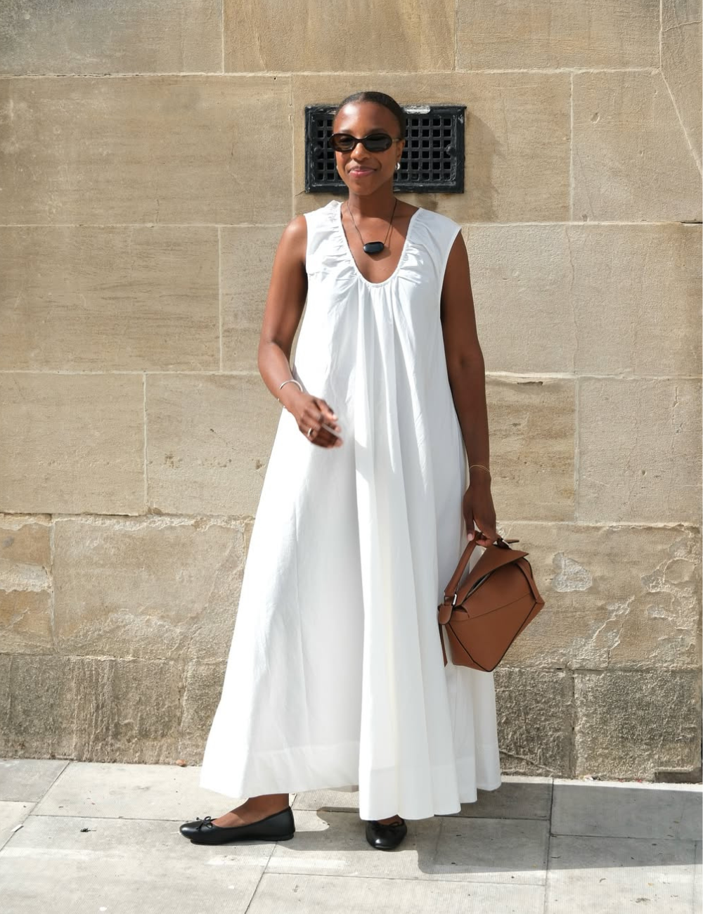 Mulher posa mostranso como se vestir bem com vestido branco fluido de algodão, bolsa marrom estruturada e sapatilhas pretas.