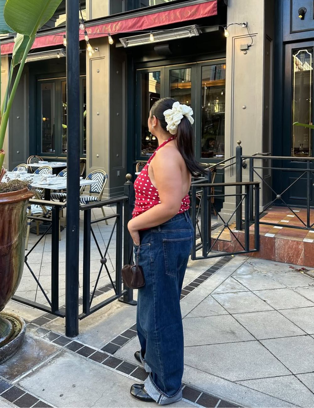 Mulher posa de perfil com blusa vermelha de alcinha e poás brancos, jeans largo e scrunchie creme, revelando estilo casual.