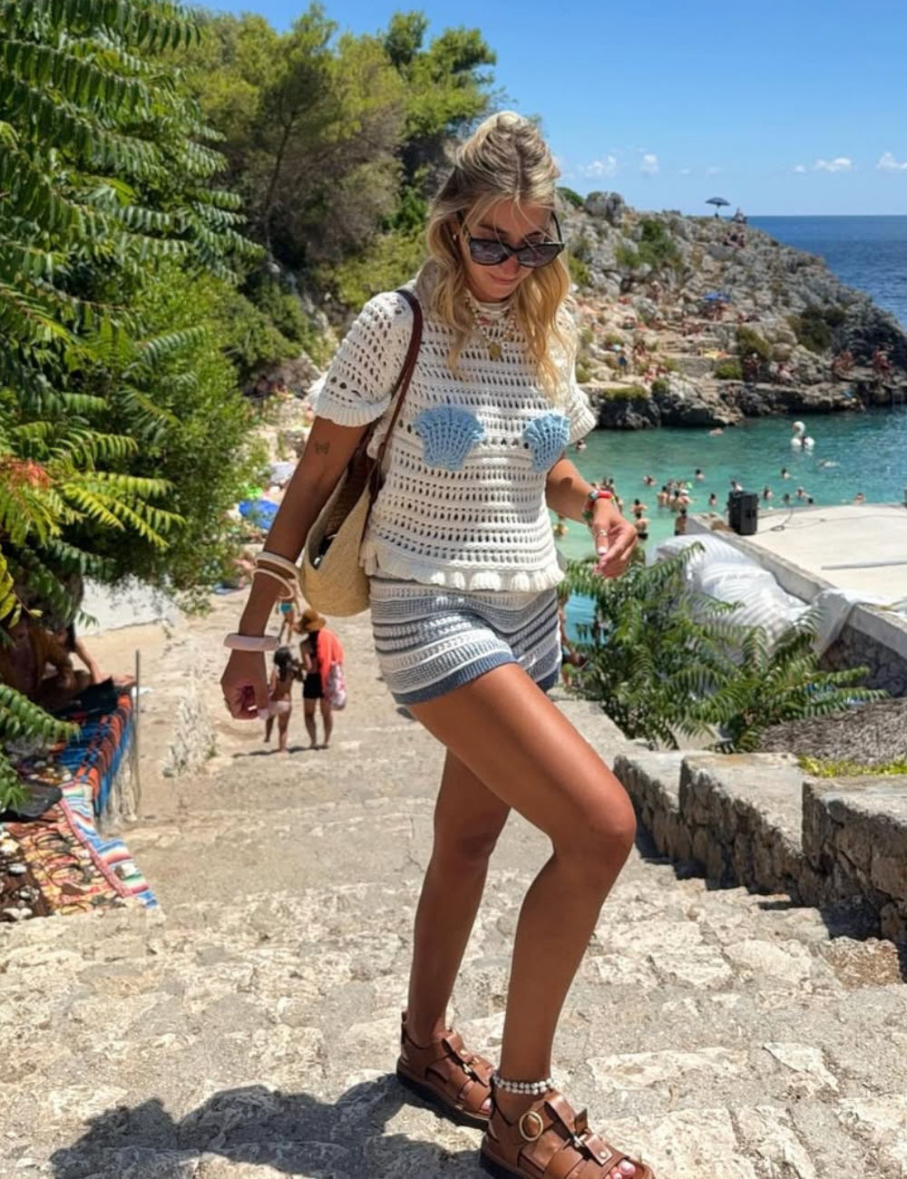 Modelo caminha em escadaria de pedra com blusa de crochê branca de manga curta e detalhes azuis, combinada com shorts.
