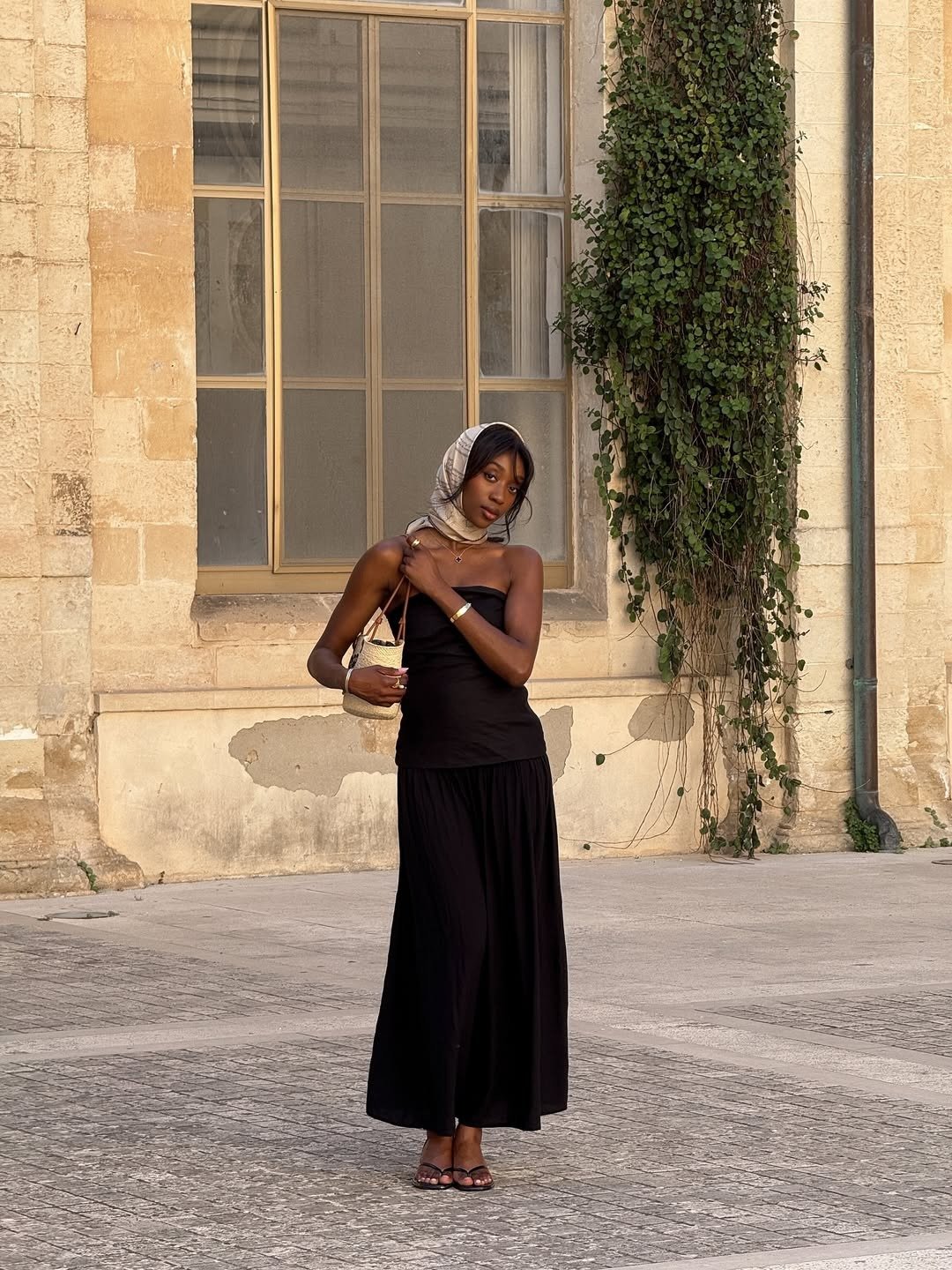 Modelo posa com vestido preto longo e tomara que caia, combinando bolsa de ráfia pequena e lenço estampado na cabeça, em