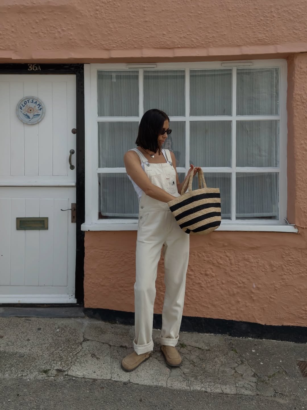 Modelo usa jardineira off-white com camiseta básica branca e bolsa de ráfia listrada preta, em clima casual e despojado,