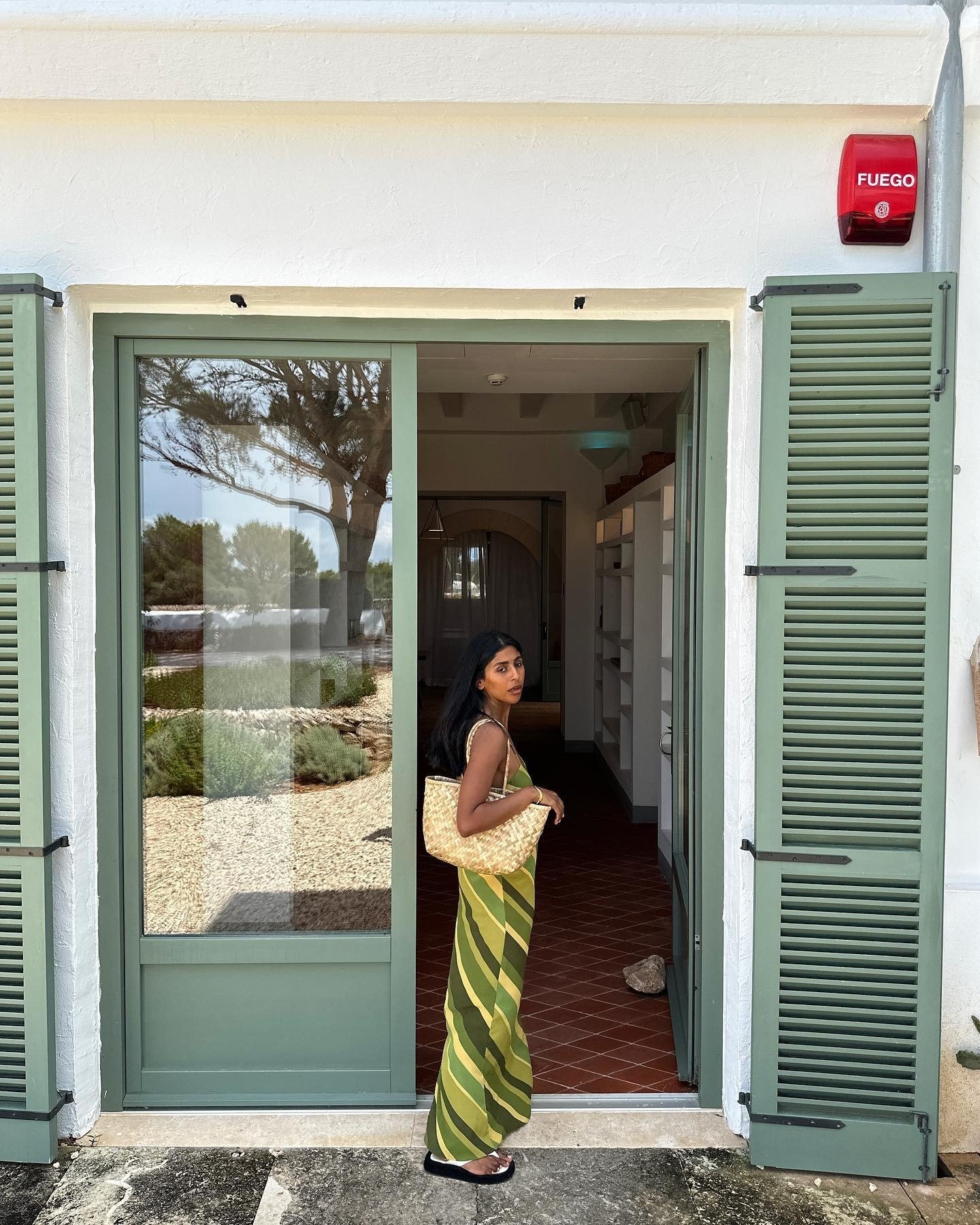 Mulher posa com vestido longo listrado verde e amarelo, usando bolsa de ráfia natural e sandálias pretas, em clima casual e