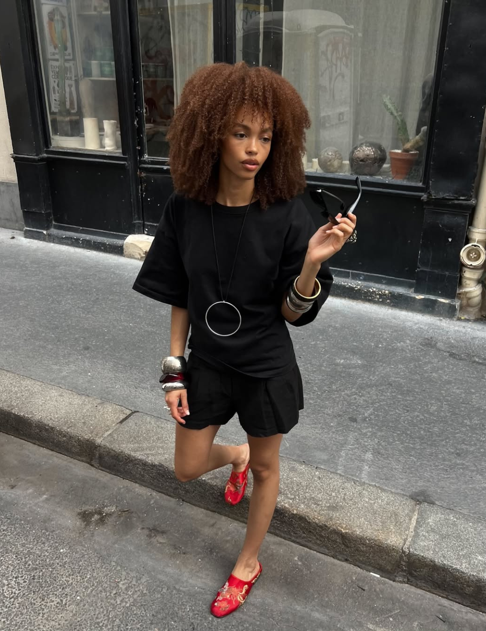 Modelo posa com look preto de camiseta solta e shorts leves, combinando acessórios metálicos e mule vermelho.