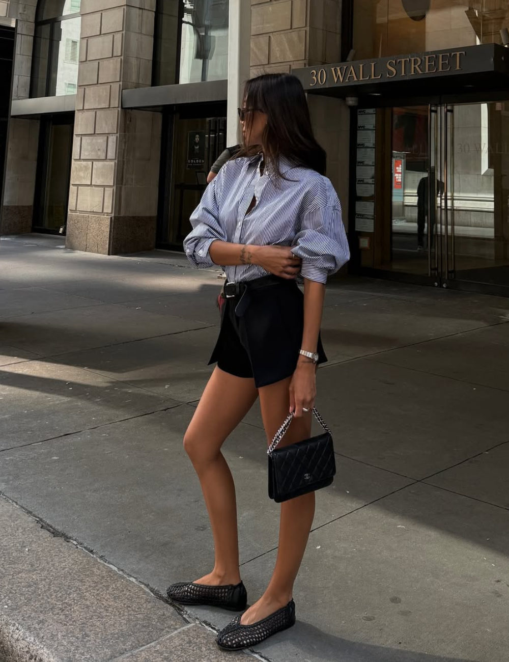 Modelo posa em shorts de alfaiataria preto com cinto, combinando camisa listrada oversized e acessórios sóbrios.