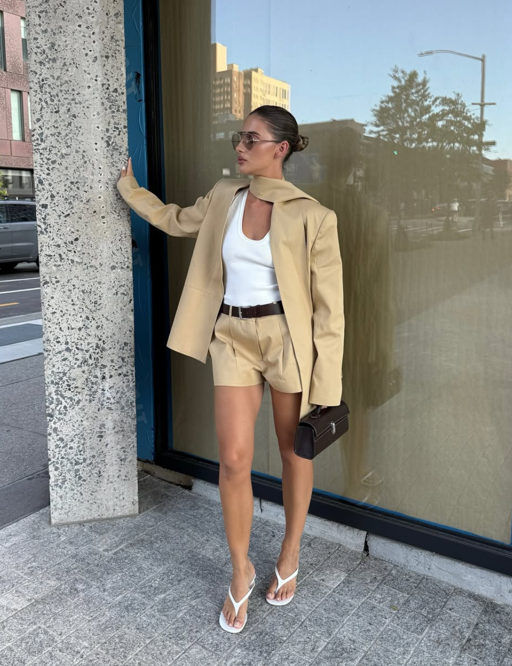Modelo posa com shorts de alfaiataria bege, blazer oversized, regata branca e sandálias minimalistas.