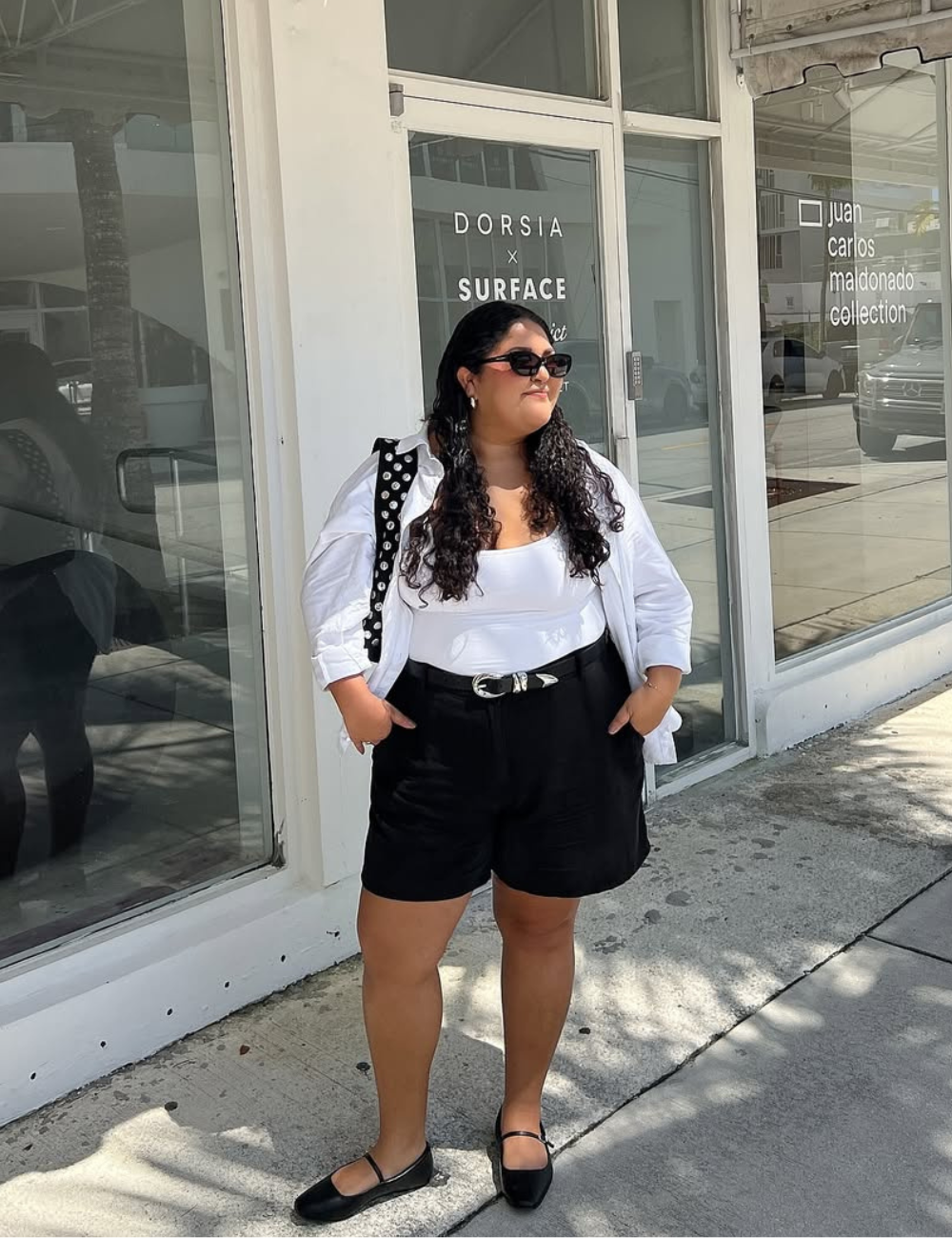 Modelo posa com shorts de alfaiataria preto de tecido estruturado, cinto com fivela prateada, regata branca, e camisa branca.