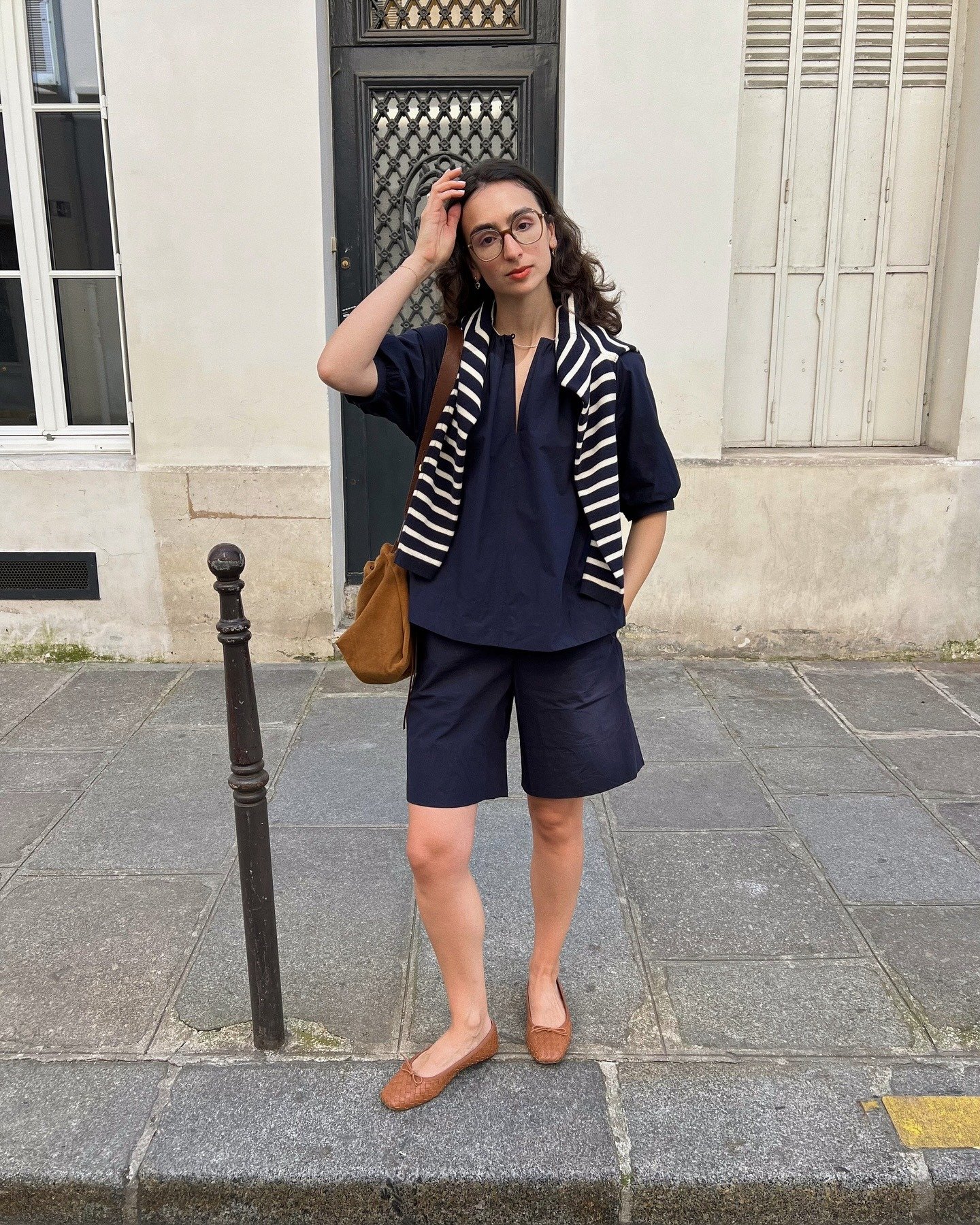 Modelo posa com look casual em tons de azul marinho, shorts e blusa soltos, com suéter listrado e acessórios caramelo,