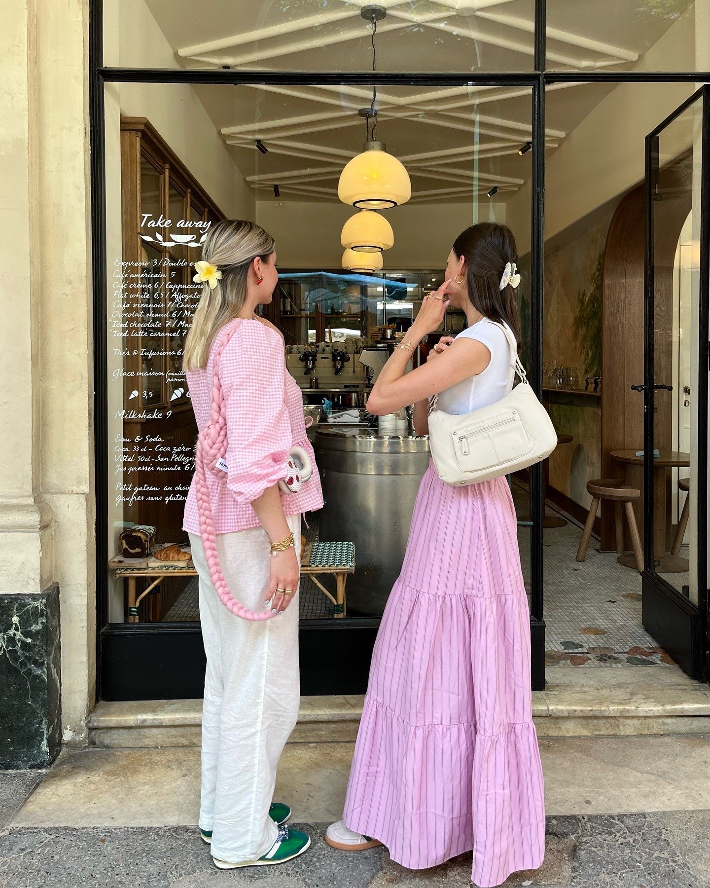 Duas mulheres posam em clima casual diante de café, usando rosa pastel e branco em blusa xadrez bufante, calça reta e saia
