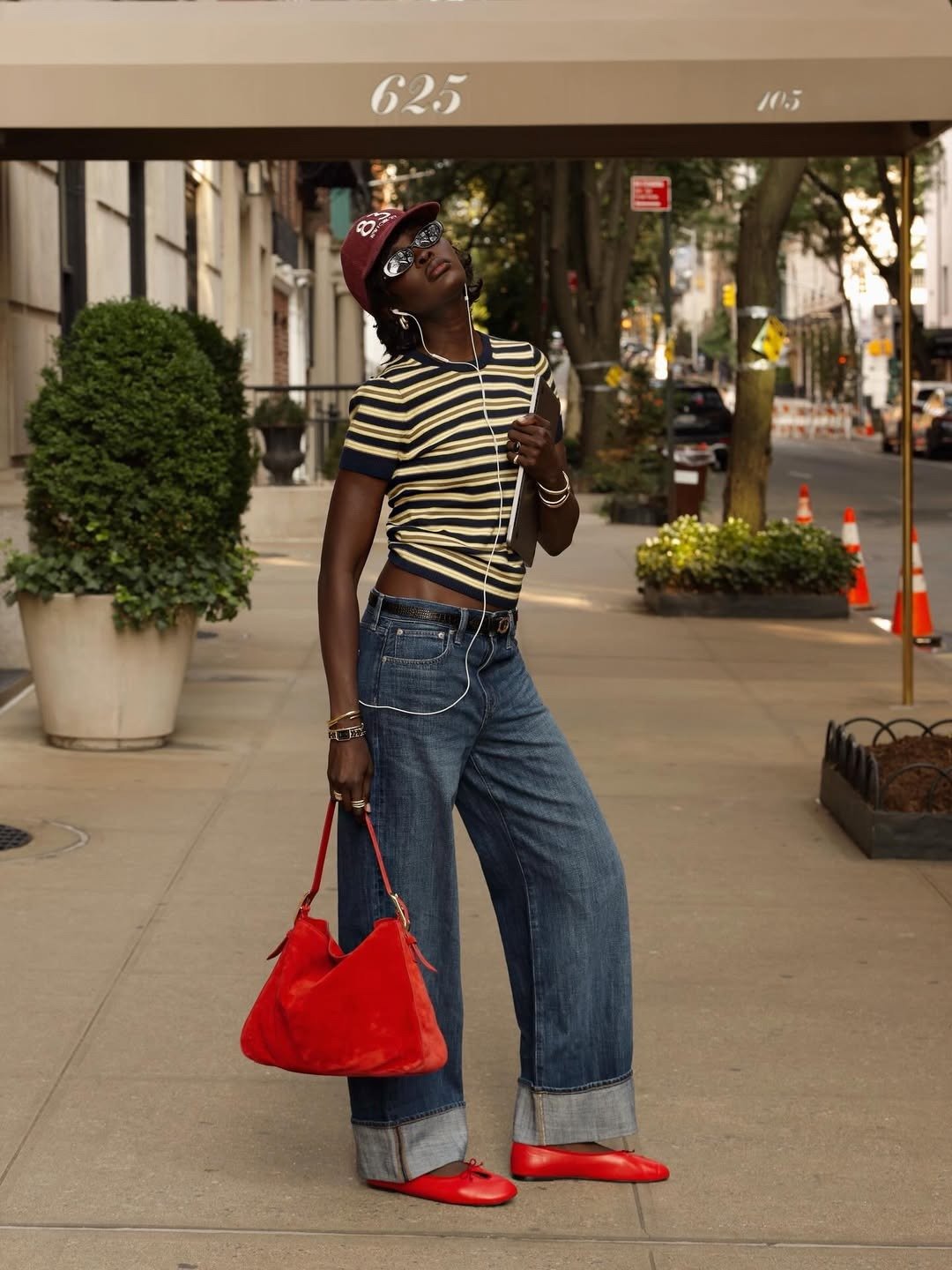 Modelo posa na rua com jeans largo dobrado, camiseta listrada amarela e azul, sapatilhas vermelhas de couro e bolsa vermelha