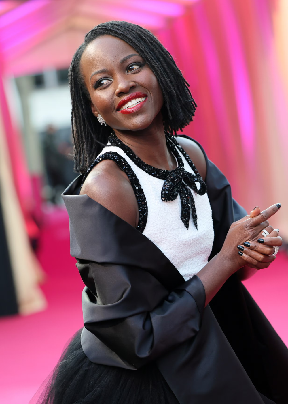 Modelo sorri olhando para o lado, exibindo corte bob em dreads pretos, vestido branco com detalhes em paetês pretos e xale.