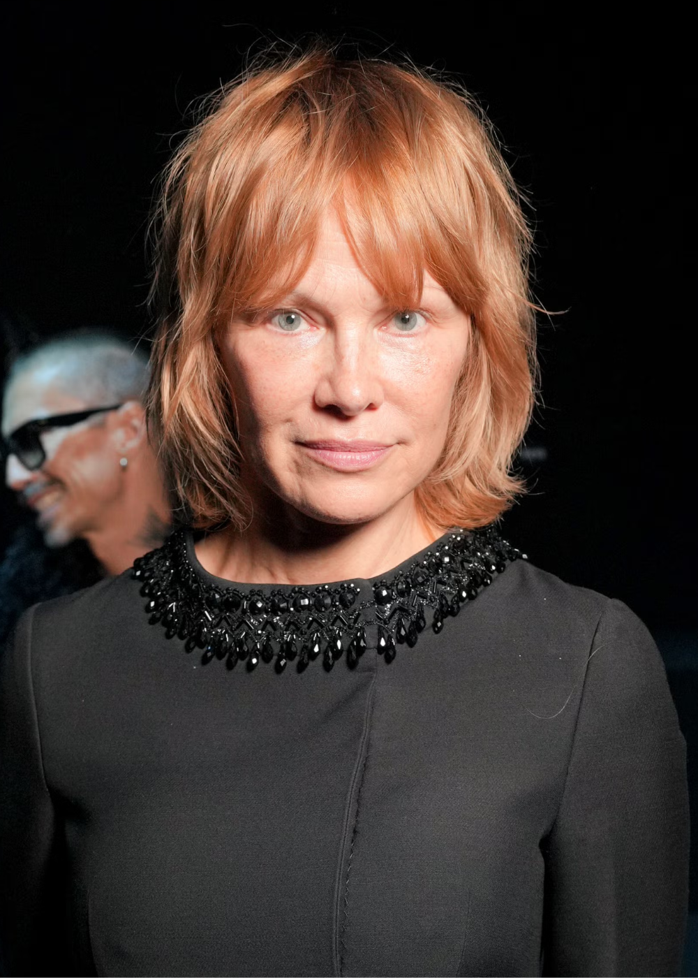 Mulher com corte bob em tom ruivo natural, vestido preto com gola bordada em pedrarias, expressão serena e confiante.