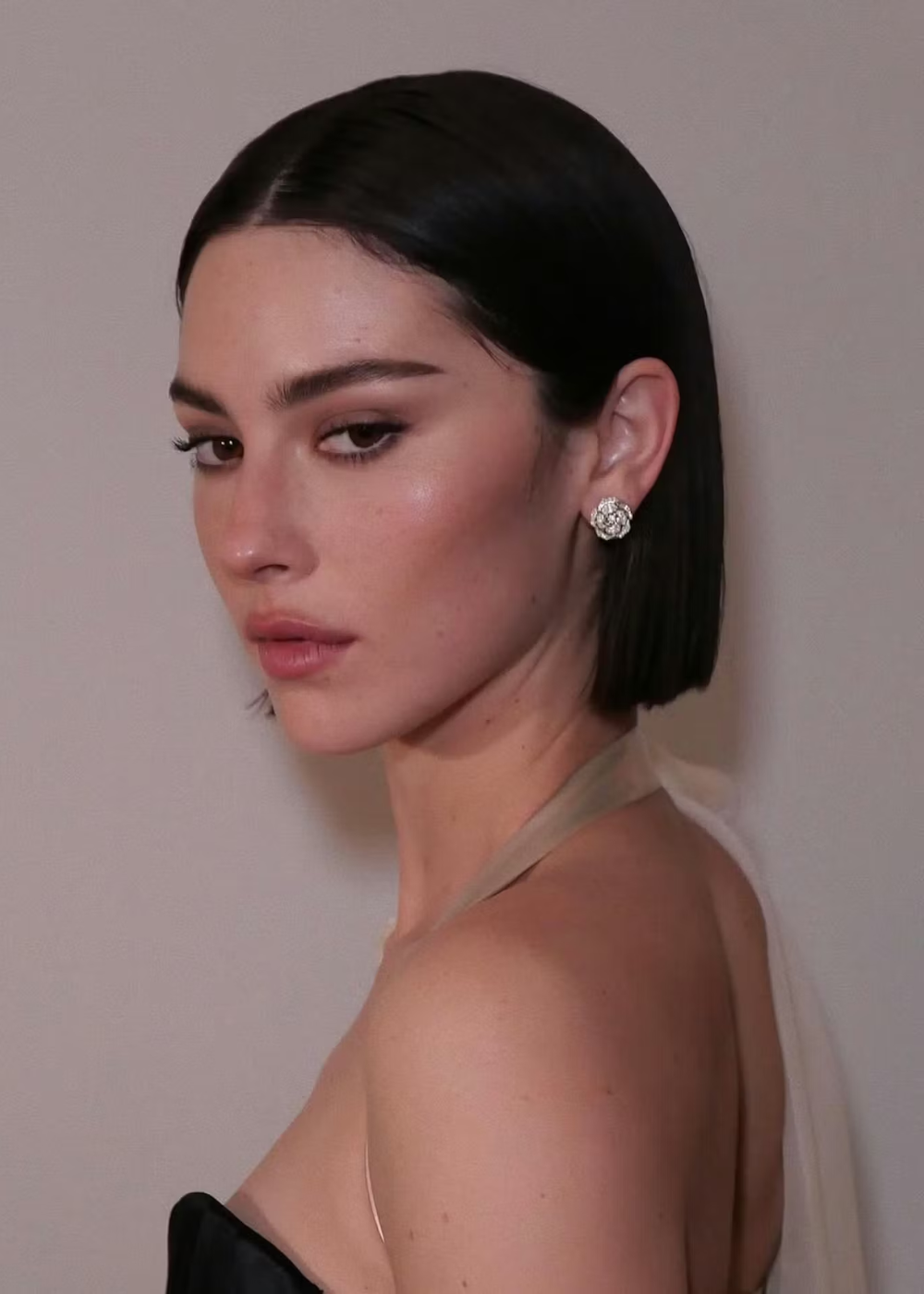 Modelo posa de perfil com corte bob liso e reto, cabelo preto brilhante, brincos florais e look sofisticado em tons neutros.