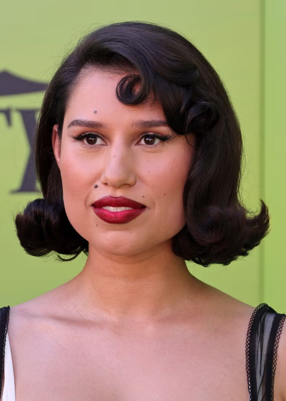 Mulher com corte bob em cabelo preto e ondulado, maquiagem marcante com batom vinho, em fundo verde vibrante.