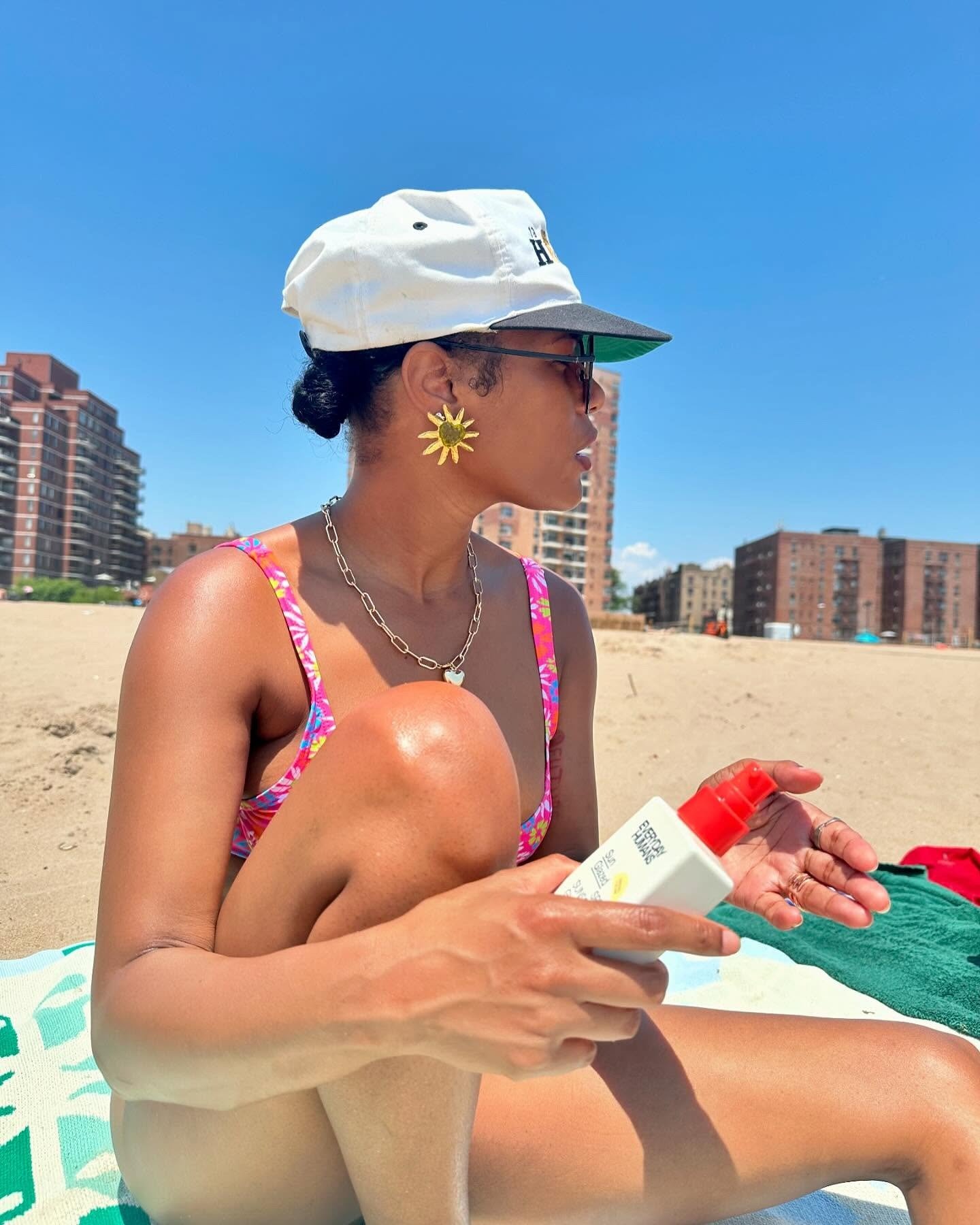 Mulher na praia com biquíni estampado rosa, boné branco e brincos dourados, cuidando da pele para evitar assadura das coxas