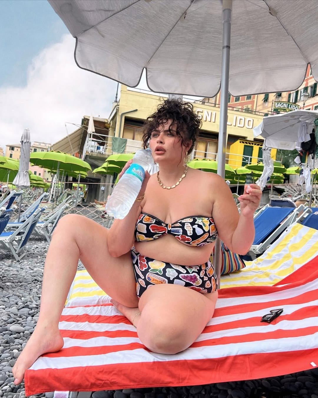 Modelo senta sob guarda-sol em praia de pedras, usando biquíni estampado com hot pants que evita assadura das coxas