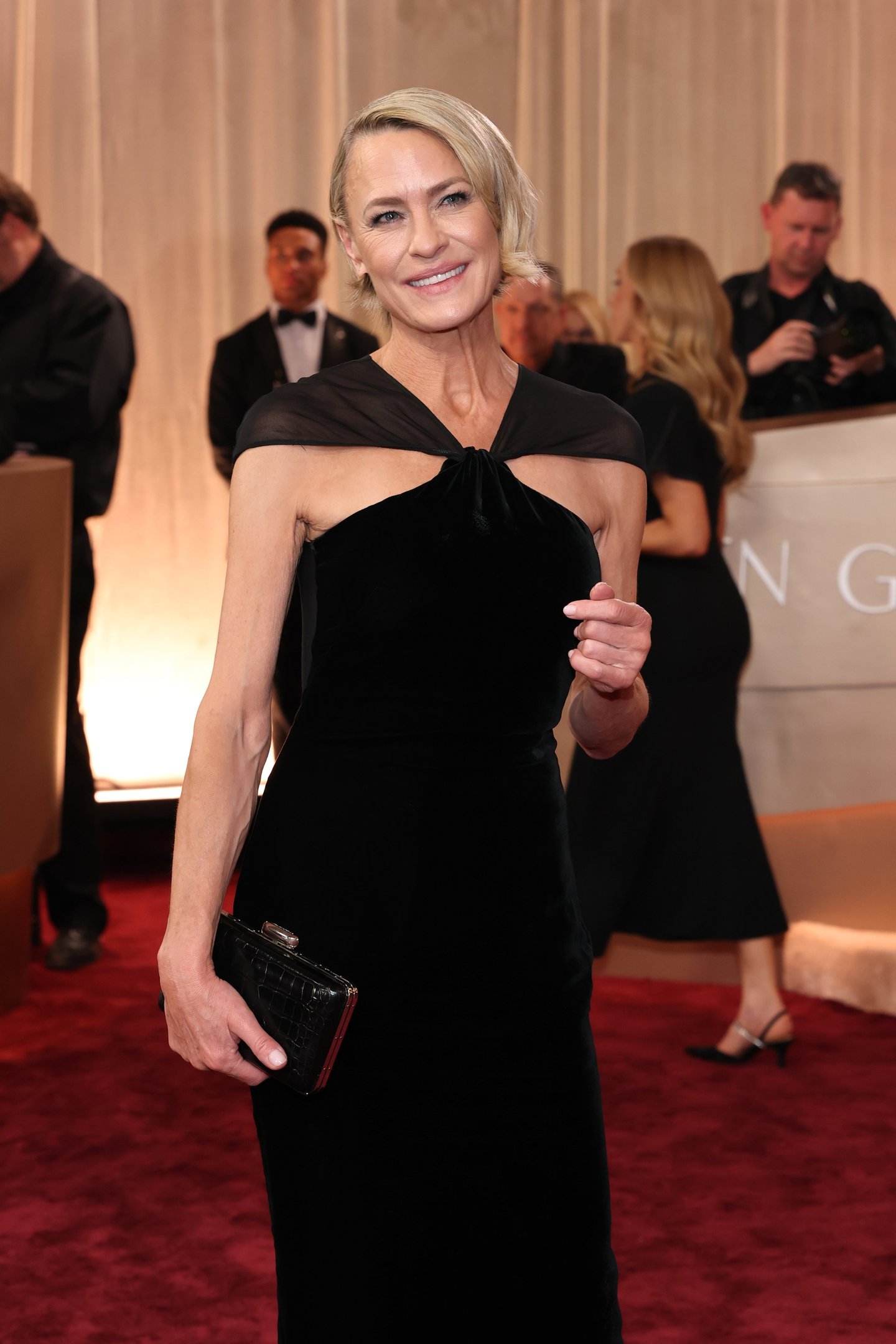 Modelo com cabelo curtinho loiro posa sorridente usando vestido preto de veludo com decote assimétrico e clutch preta