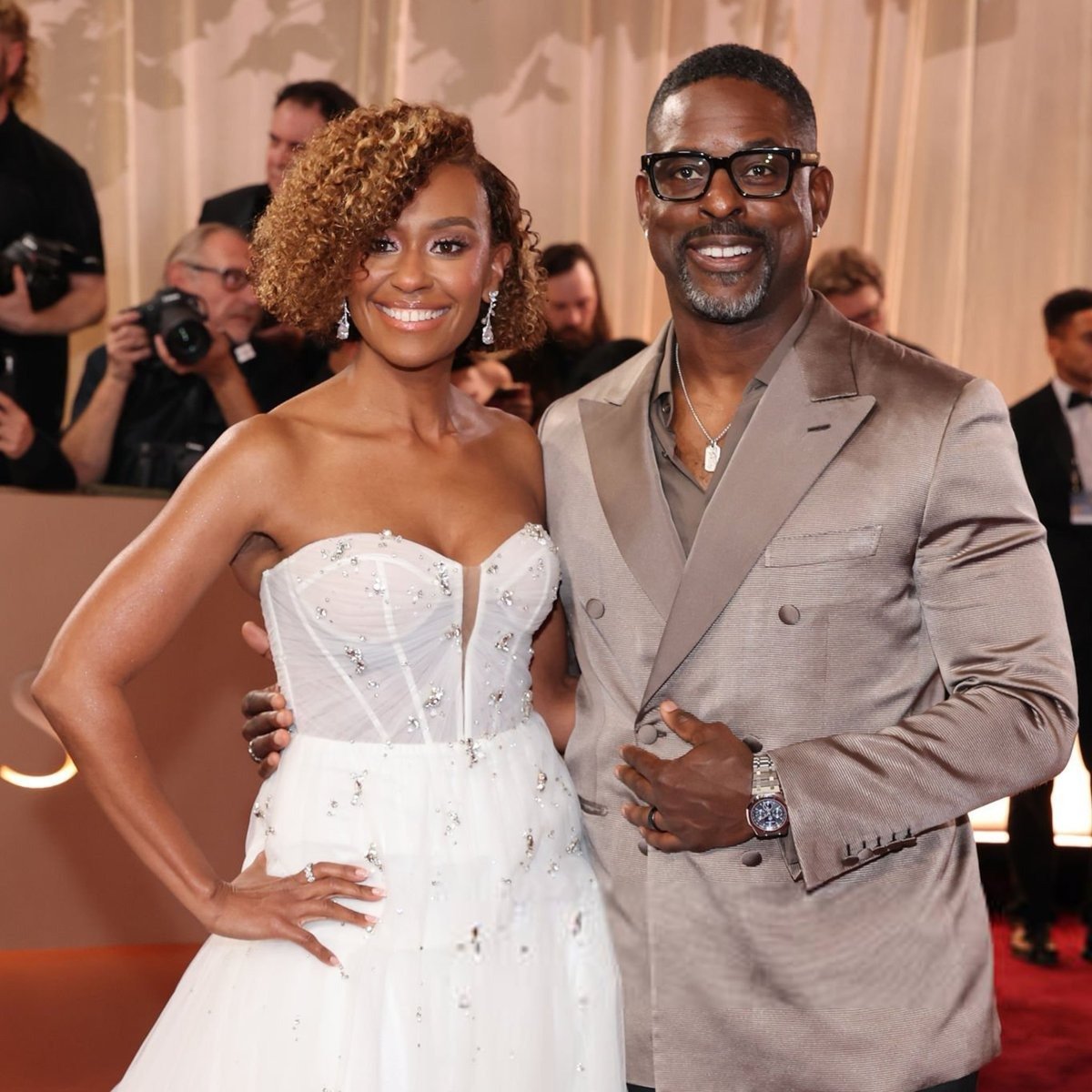 Mulher com cabelo curtinho cacheado em vestido branco bordado, posa sorrindo ao lado de homem com terno bege e óculos