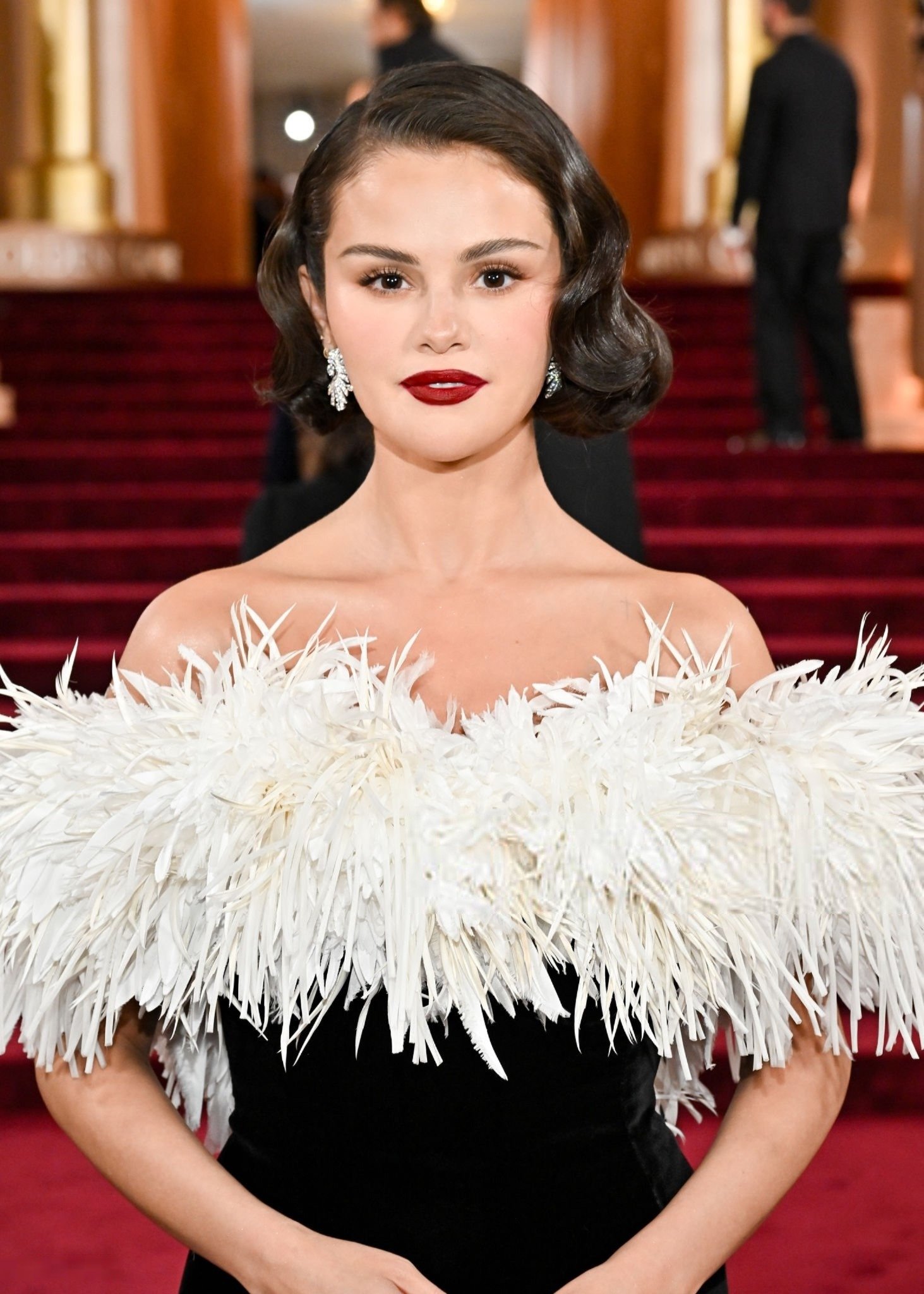 Modelo posa com cabelo curtinho ondulado, vestido preto de veludo com gola branca de penas, batom vinho e brincos