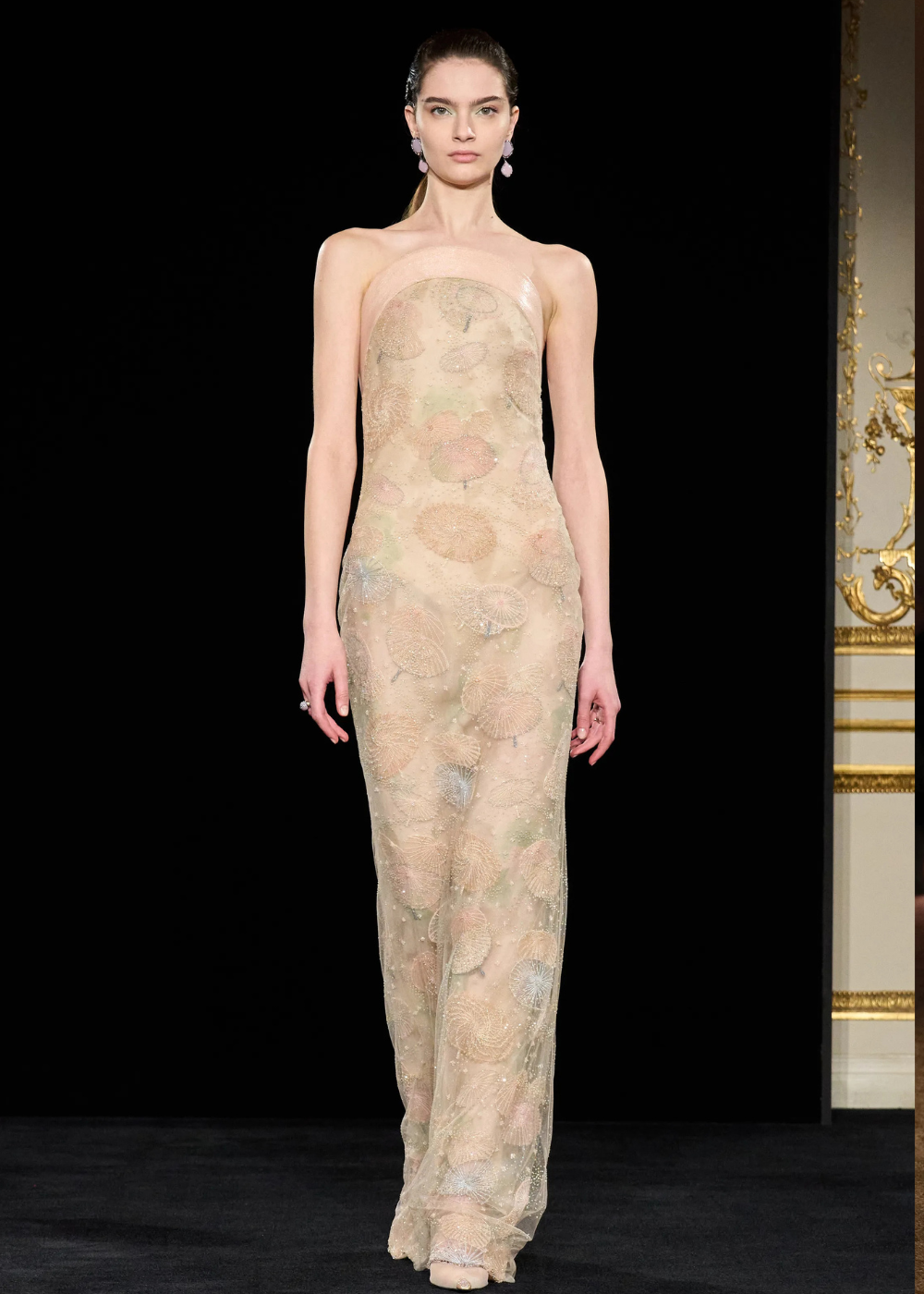 Modelo posa com vestido longo tomara que caia em tule nude bordado com texturas delicadas em tons pastel, refletindo as