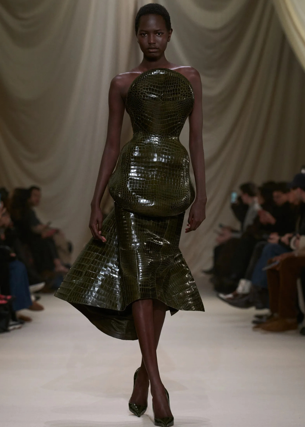 Modelo caminha com postura firme e confiante em vestido estruturado verde musgo de alta-costura, textura croco brilhante e