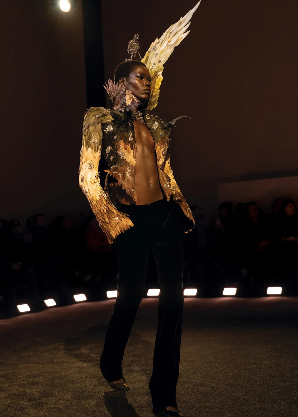 Modelo posa com jaqueta de alta-costura em penas douradas e marrons, combinada a calça preta, evidenciando tendências da