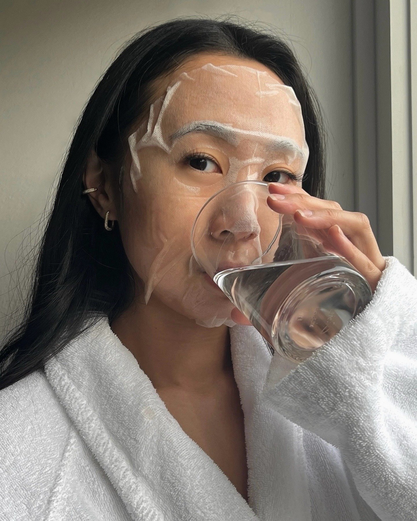Mulher com máscara facial de tecido transparente e roupão branco bebe água em ambiente neutro, expressando cuidado e detox