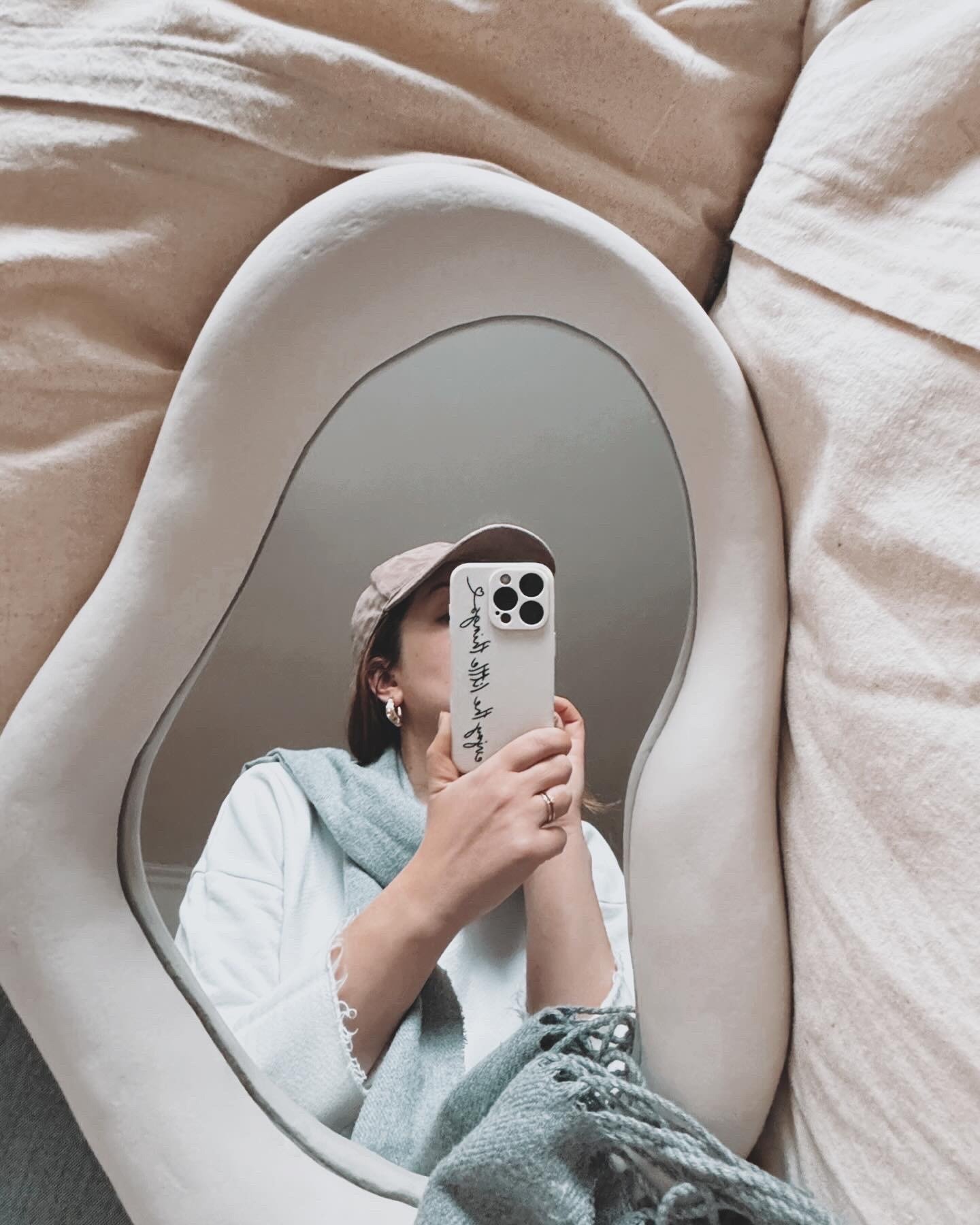 Espelho em casa com borda orgânica em tons neutros reflete mulher com casaco branco, lenço cinza e boné bege, clima