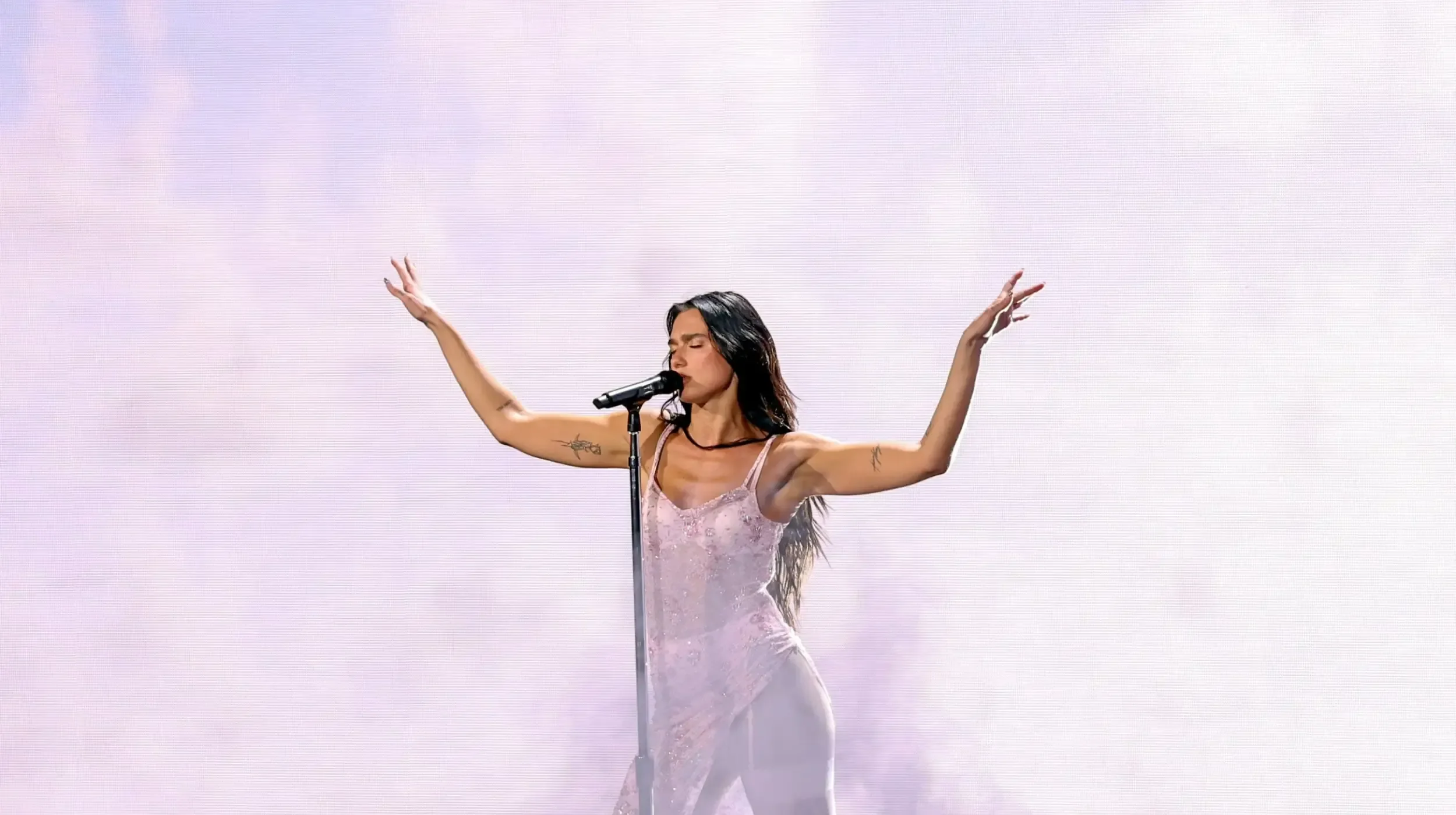 Modelo canta com braços definidos erguidos em vestido rosa claro translúcido de tecido leve e ambiente etéreo em fundo lilás